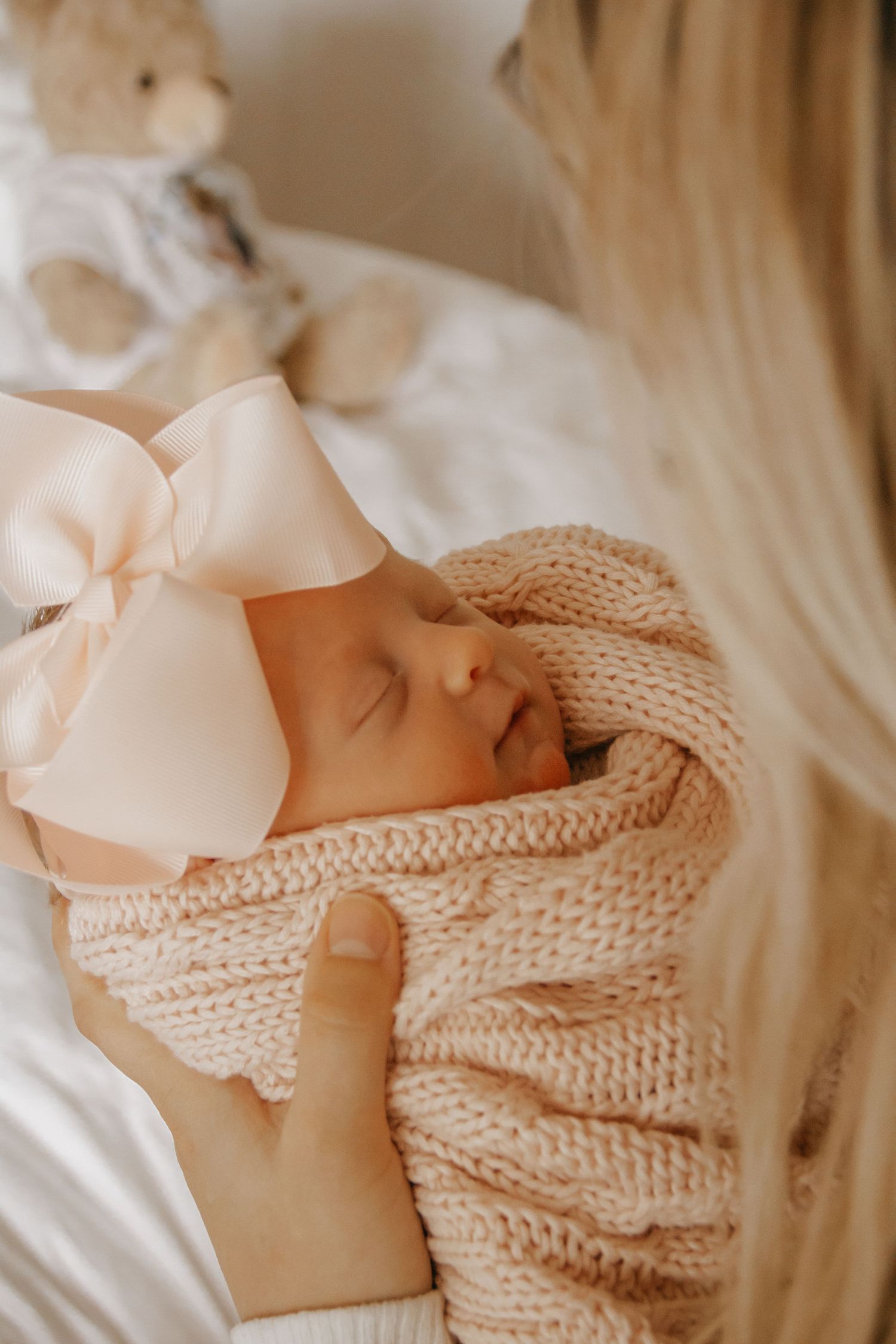 Sleeping newborn baby wrapped in a soft pink knit blanket with a large bow headband, cradled in mother's arms.