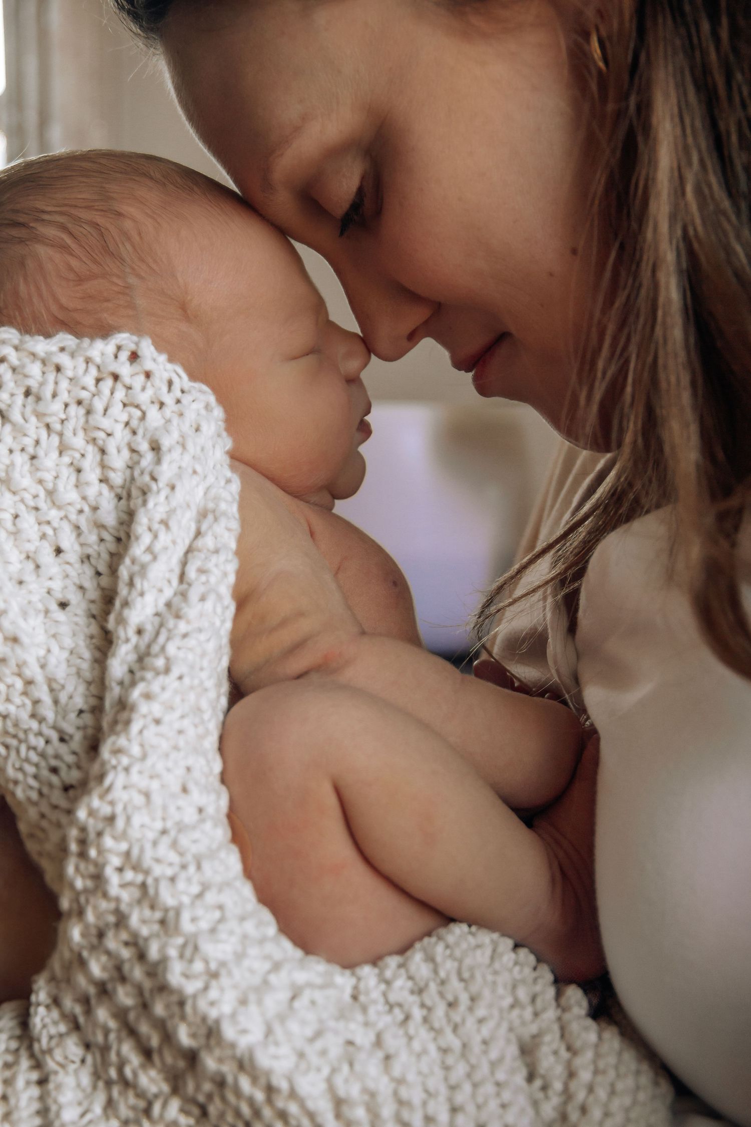 A tender moment of a mother holding her newborn baby wrapped in a white knit blanket, touching foreheads gently.