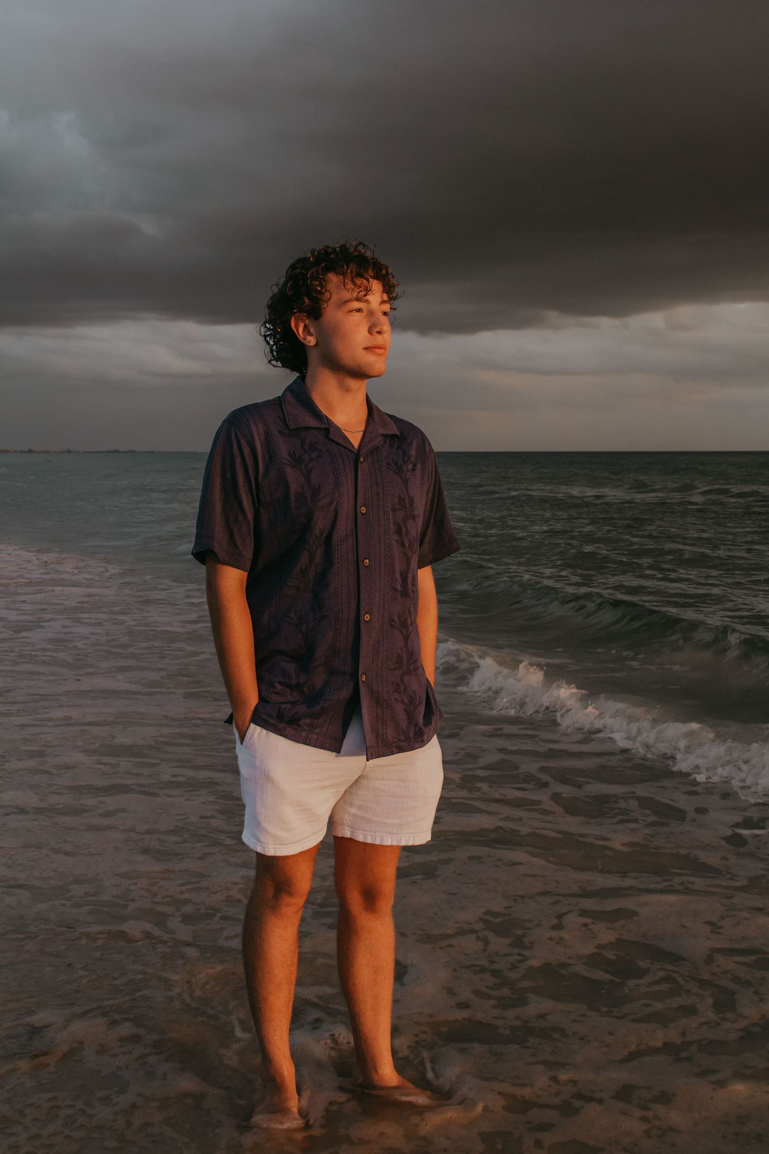 Young man with curly hair stands barefoot on a sandy beach in white shorts and dark shirt under dramatic stormy skies.