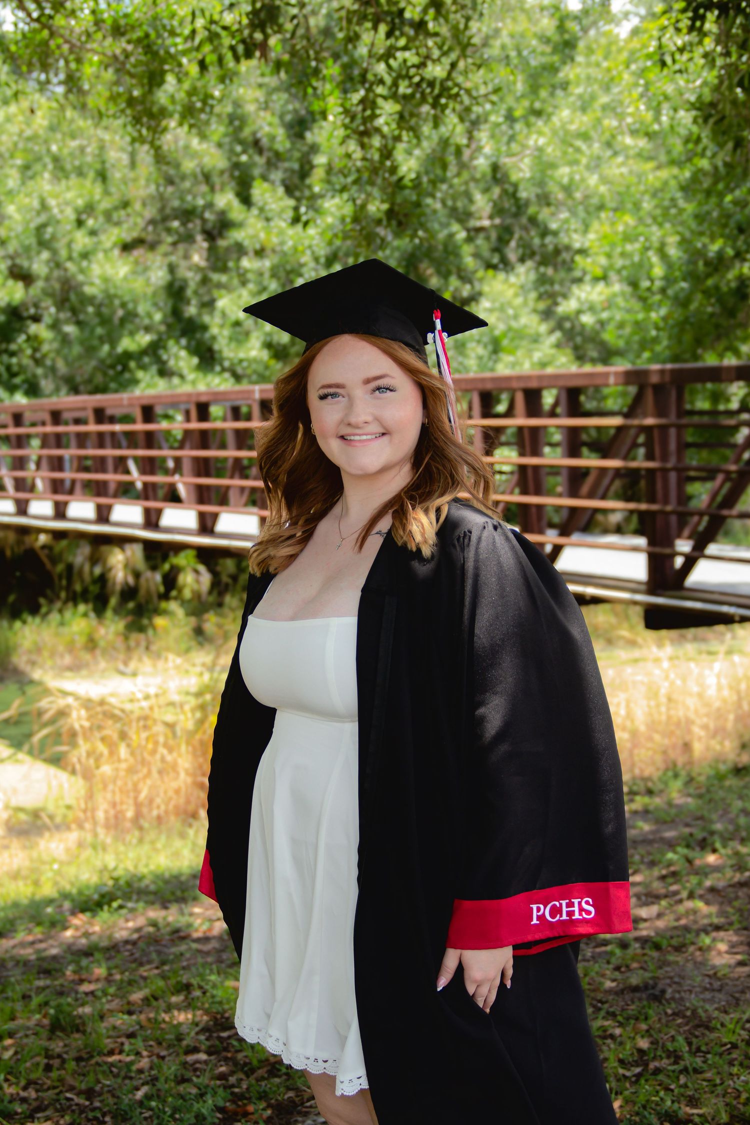 Smiling graduate in black cap and gown with PCHS trim over white dress, posing outdoors near a wooden bridge.