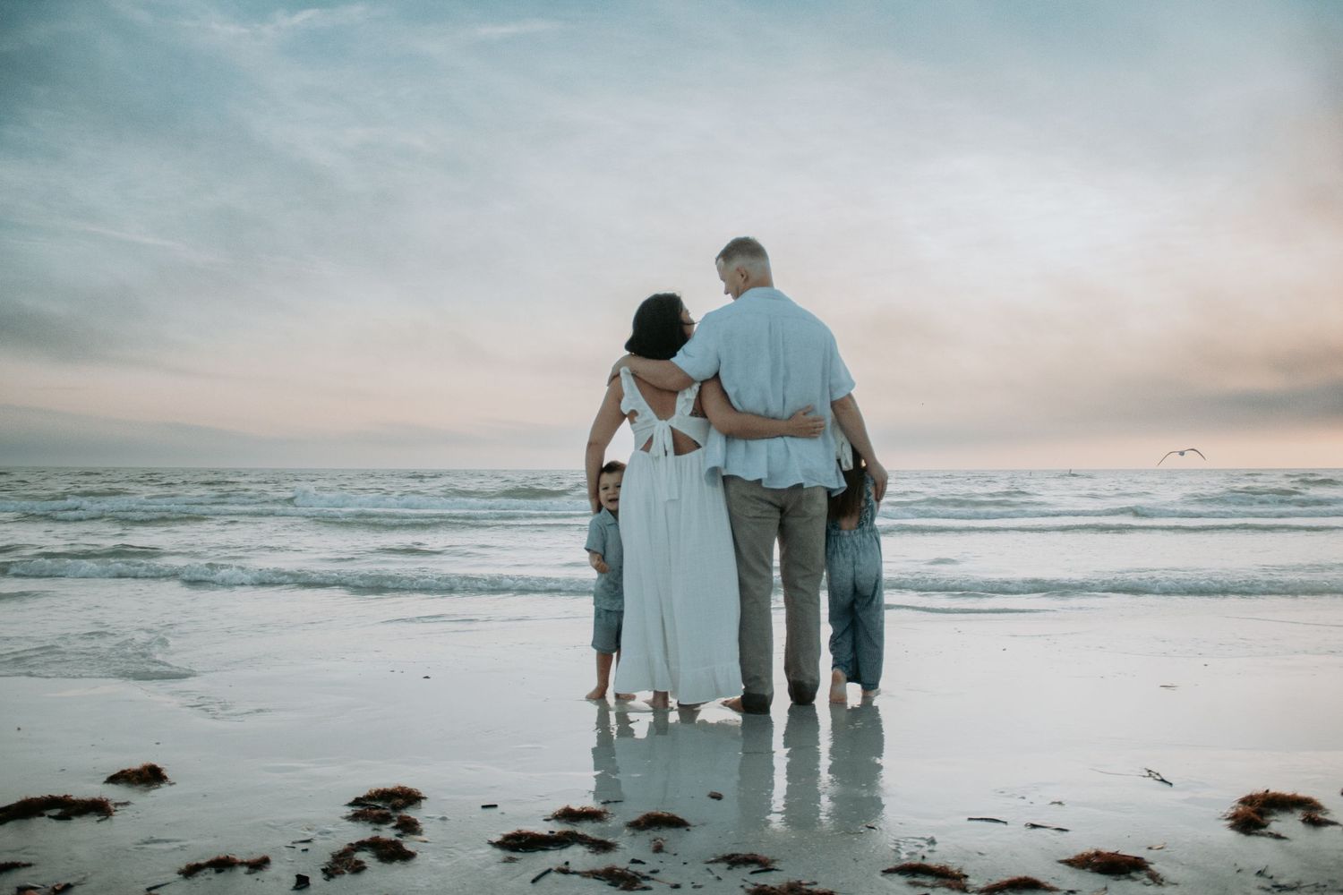 Family of four standing together on a beach at sunset, viewed from behind, waves gently lapping the shore.