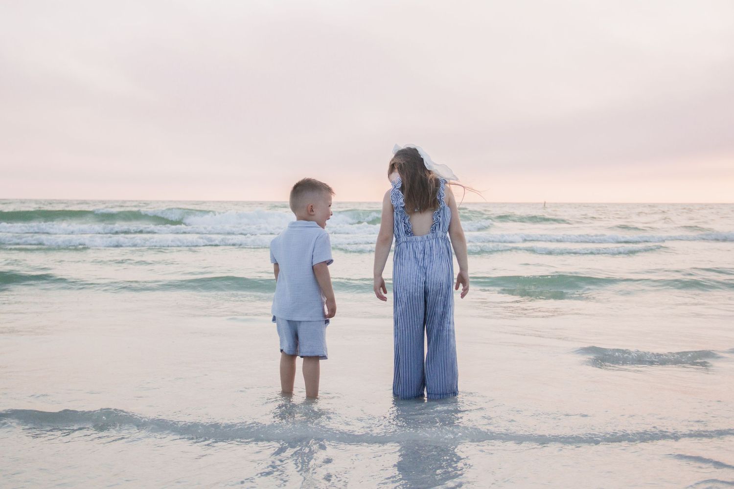 Two children standing in ocean waves at sunset on a sandy beach, wearing light blue outfits.