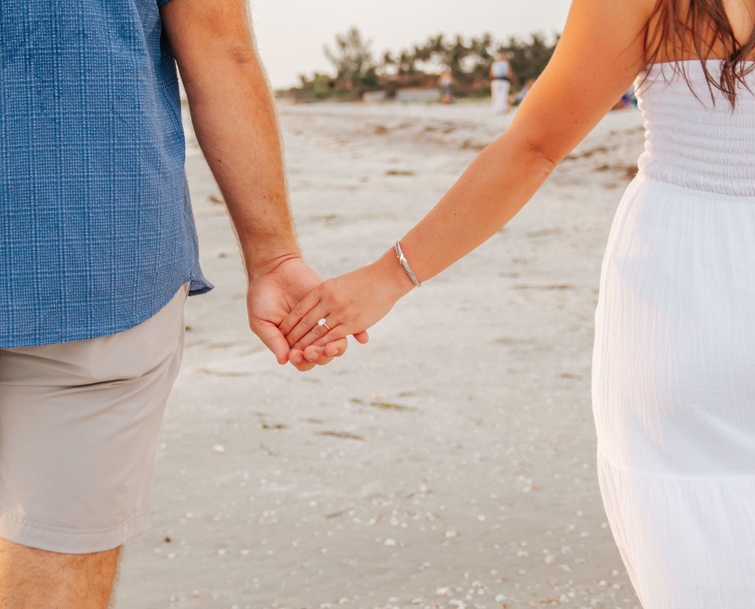 Couple holding hands on a sandy beach, woman in white dress and man in blue shirt and khaki shorts.