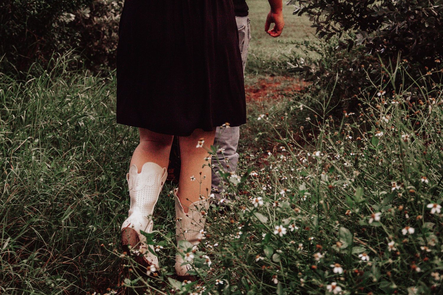 Person in black dress and white cowboy boots walking through a wildflower meadow.