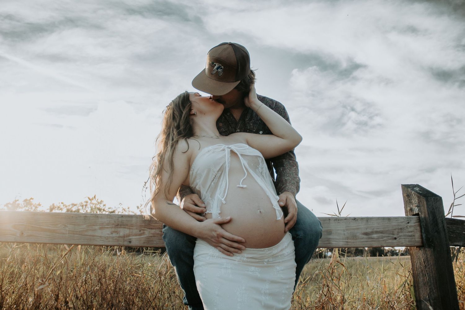Couple sharing a tender kiss during outdoor maternity photoshoot near a wooden fence in a golden field.