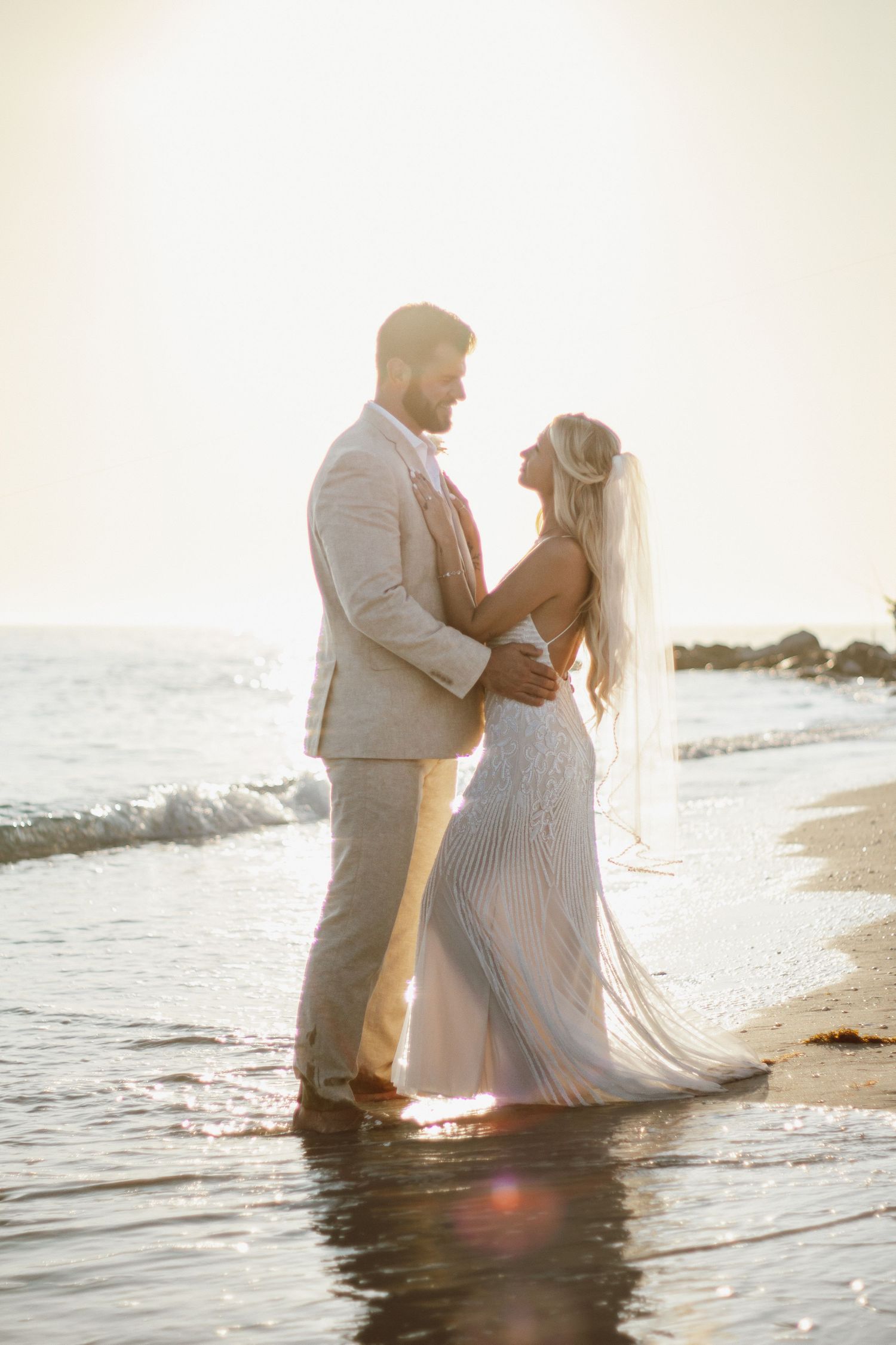 Bride and groom embrace on a sunlit beach, waves gently washing ashore, in elegant wedding attire at golden hour.