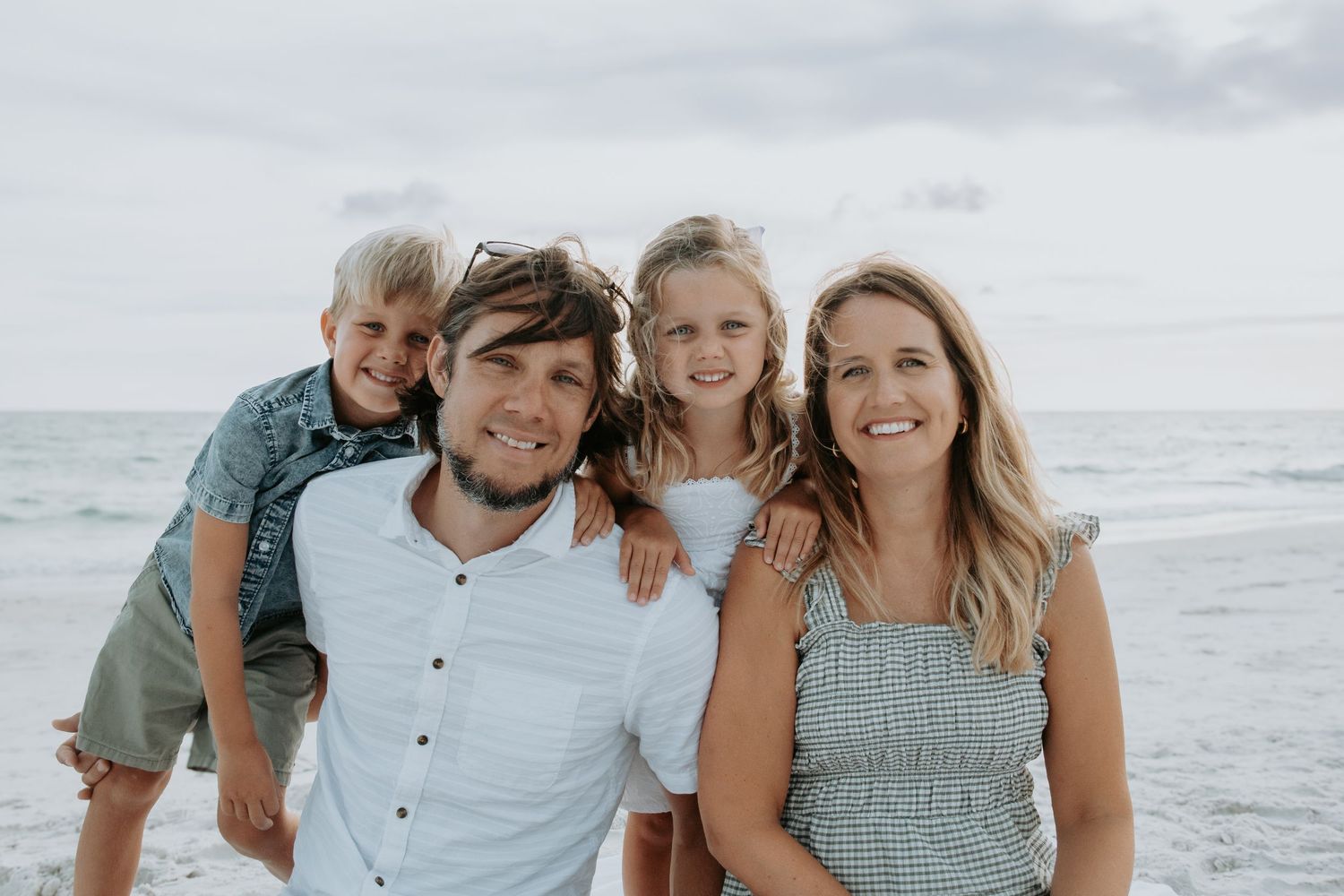 Happy family of four smiling together on a beach, with ocean waves in the background on a cloudy day.