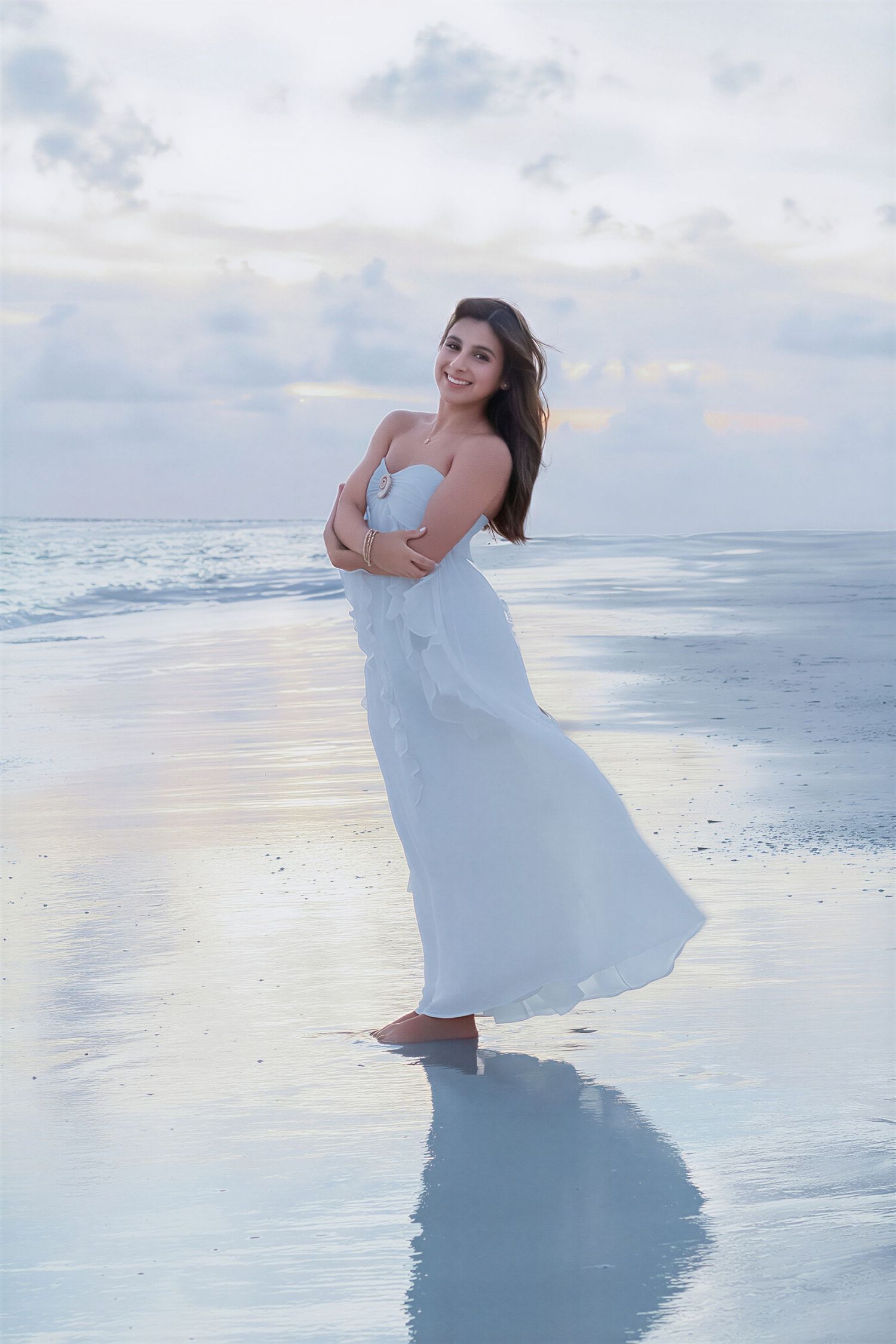 Young woman in white maxi dress standing barefoot on a serene beach at dusk with gentle waves.