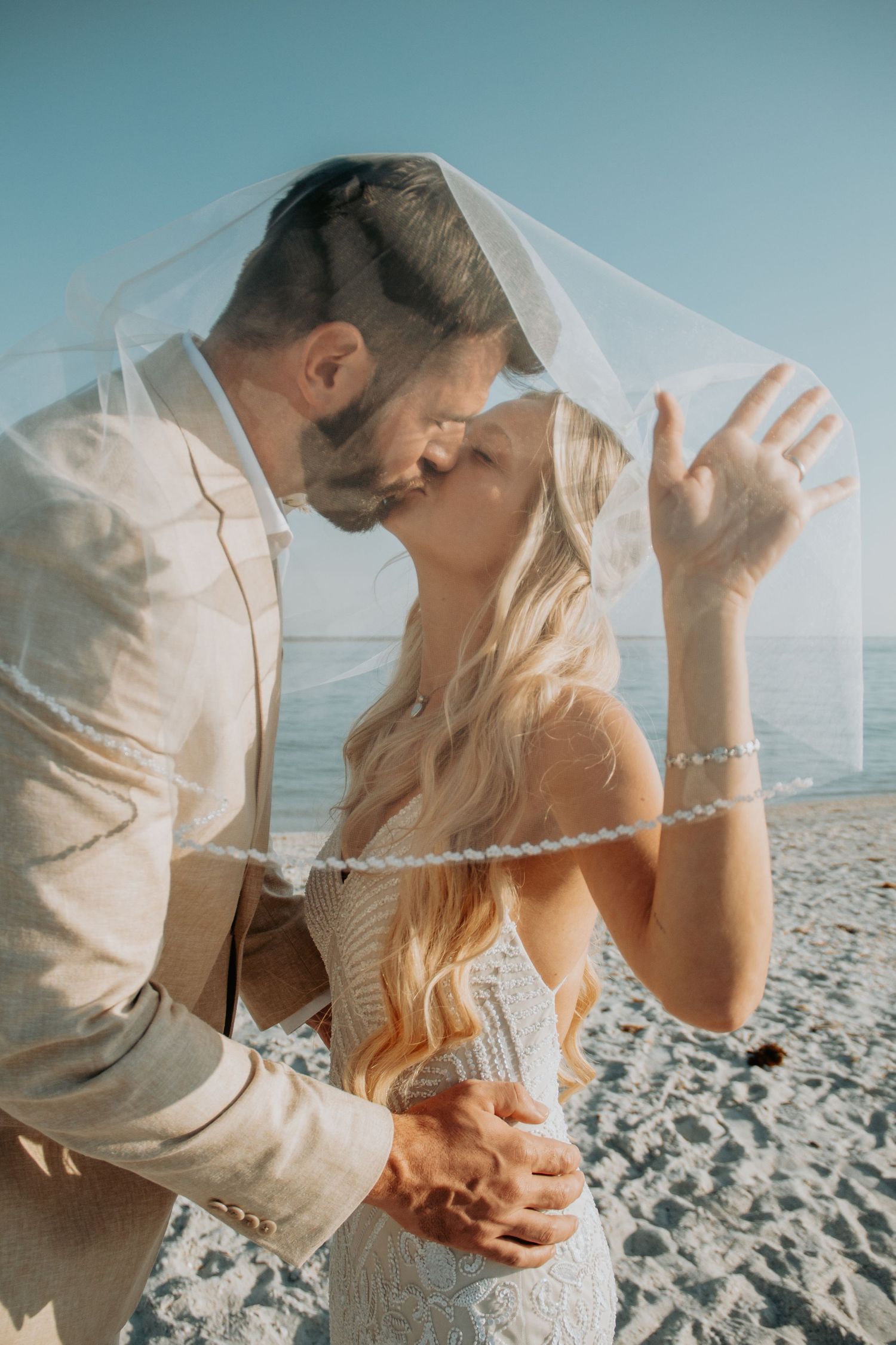 Bride and groom sharing a romantic kiss under a veil on a sunny beach wedding day.