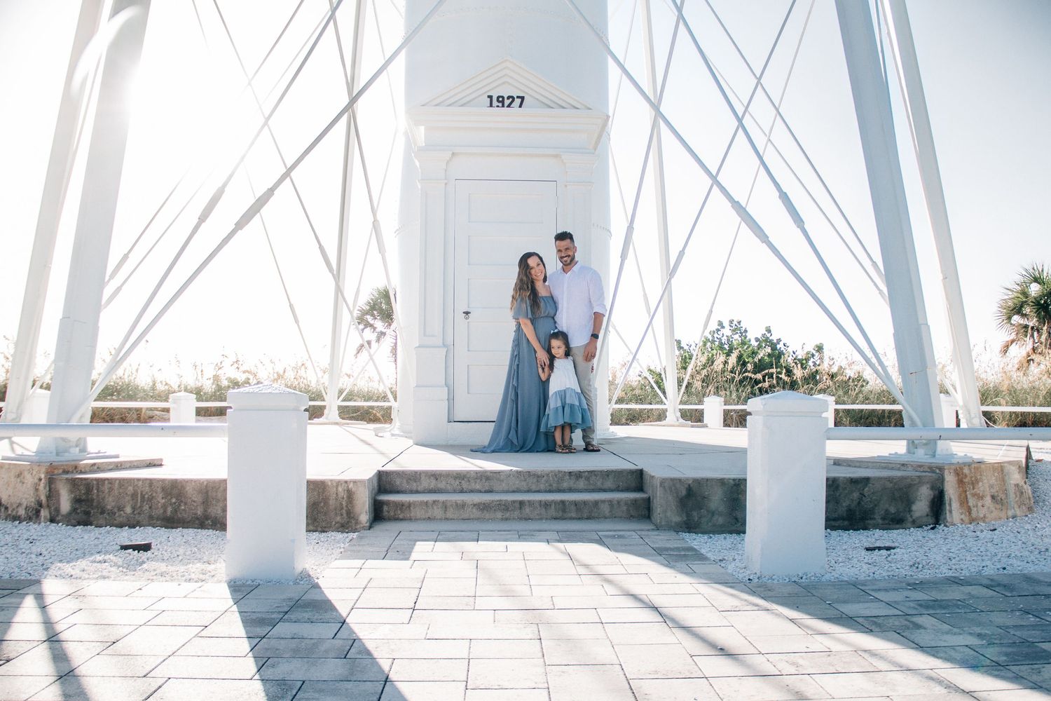 Family of three posing at a white lighthouse entrance with geometric architectural details on a sunny day.