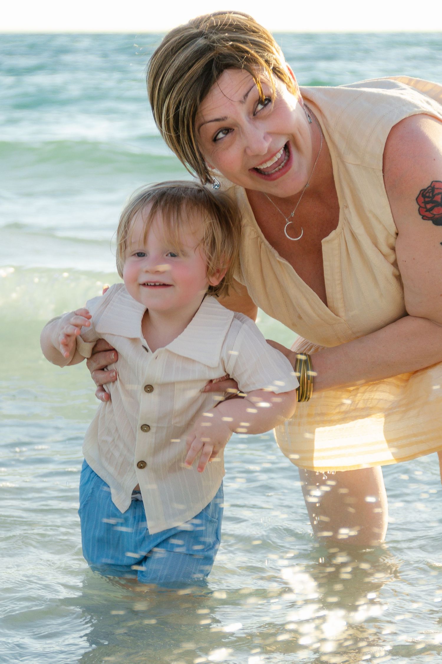 A smiling woman helps a toddler boy stand in shallow ocean waves at the beach on a sunny day.