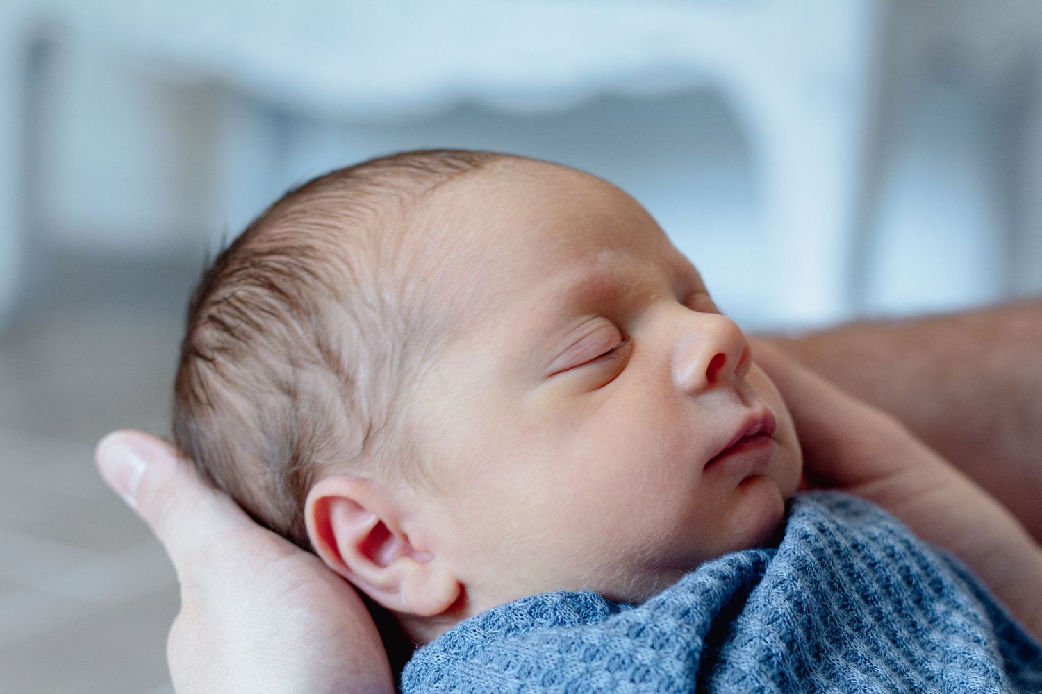 Peaceful newborn baby sleeping in an adult's hand, wrapped in a soft blue blanket, with delicate features.