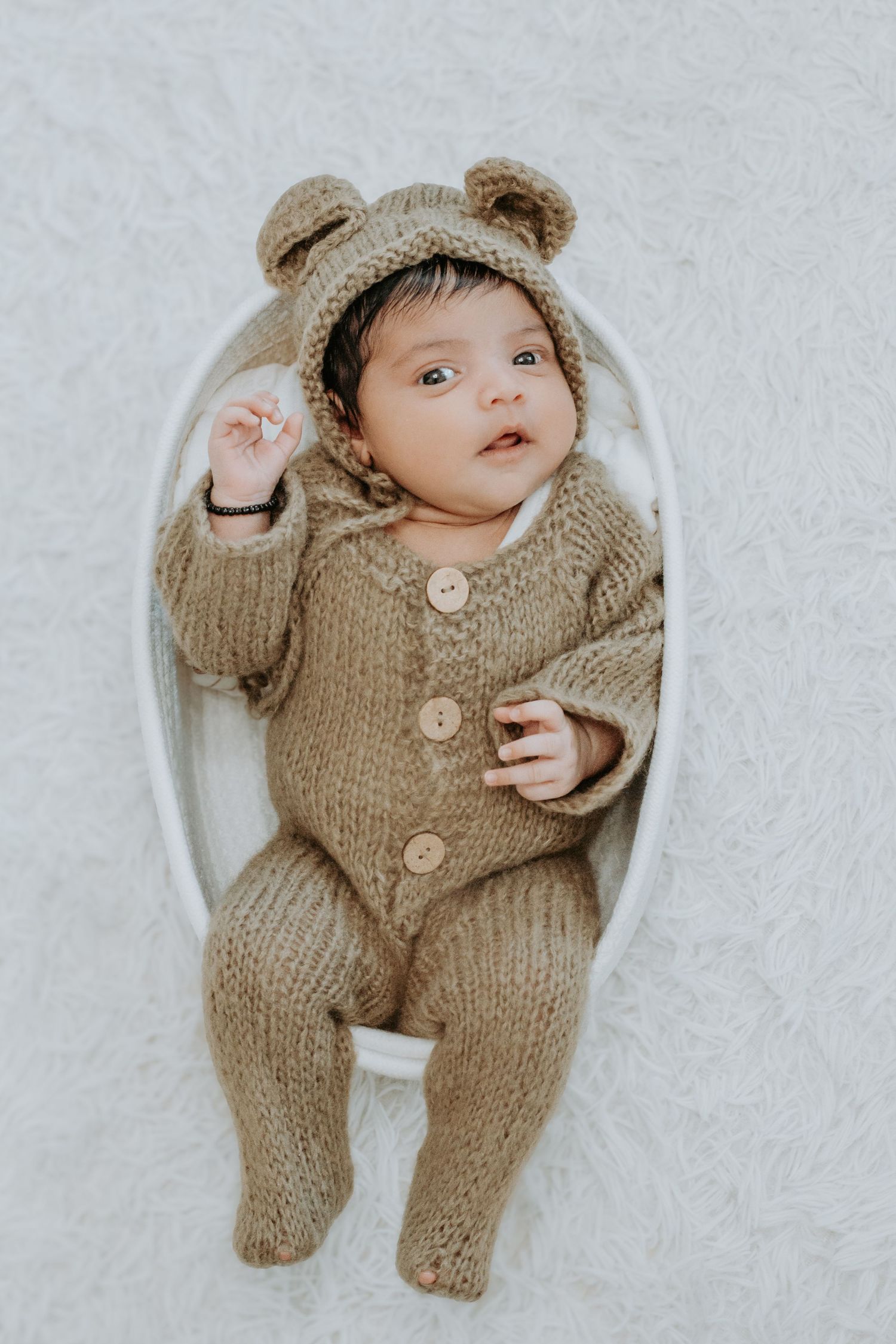 Newborn baby in a knitted bear onesie with ears hood lying in a white oval basket on a soft white background.