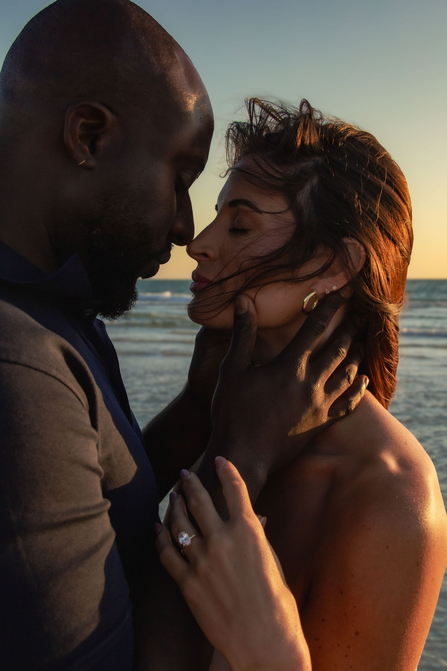 Interracial couple sharing a romantic moment at sunset on the beach, woman wearing an engagement ring.