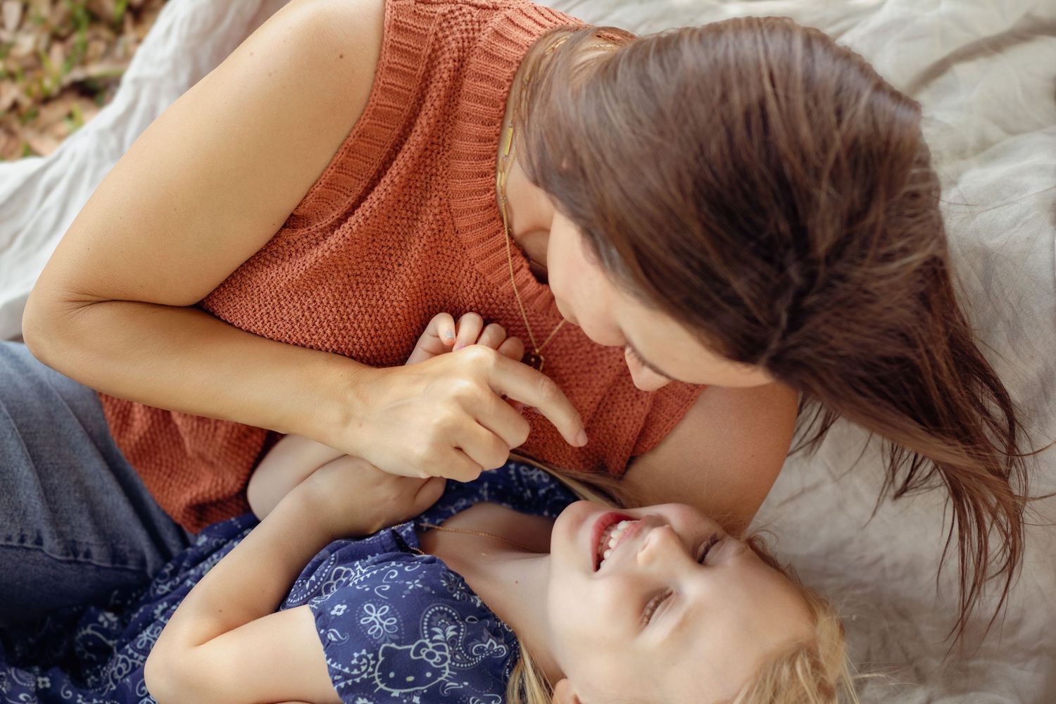 A mother and young child share a joyful moment laughing together while lying on a blanket outdoors.