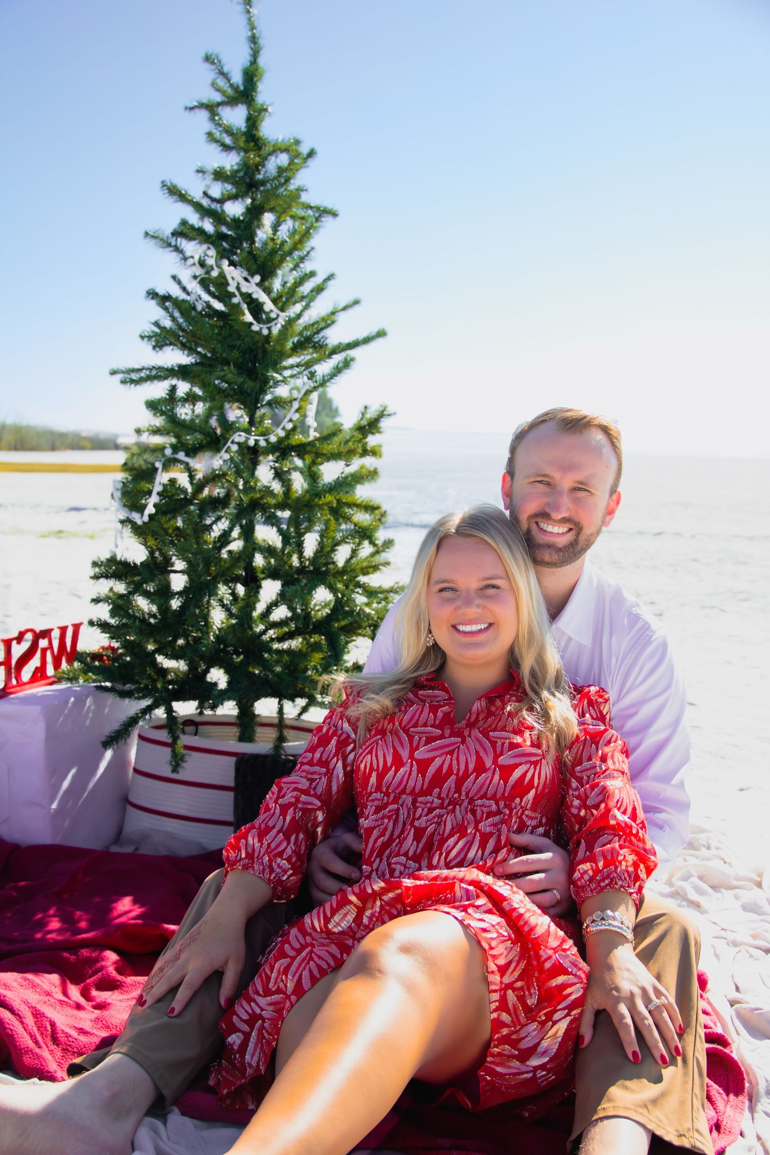 A couple sits on a beach blanket next to a decorated Christmas tree with the ocean in the background.
