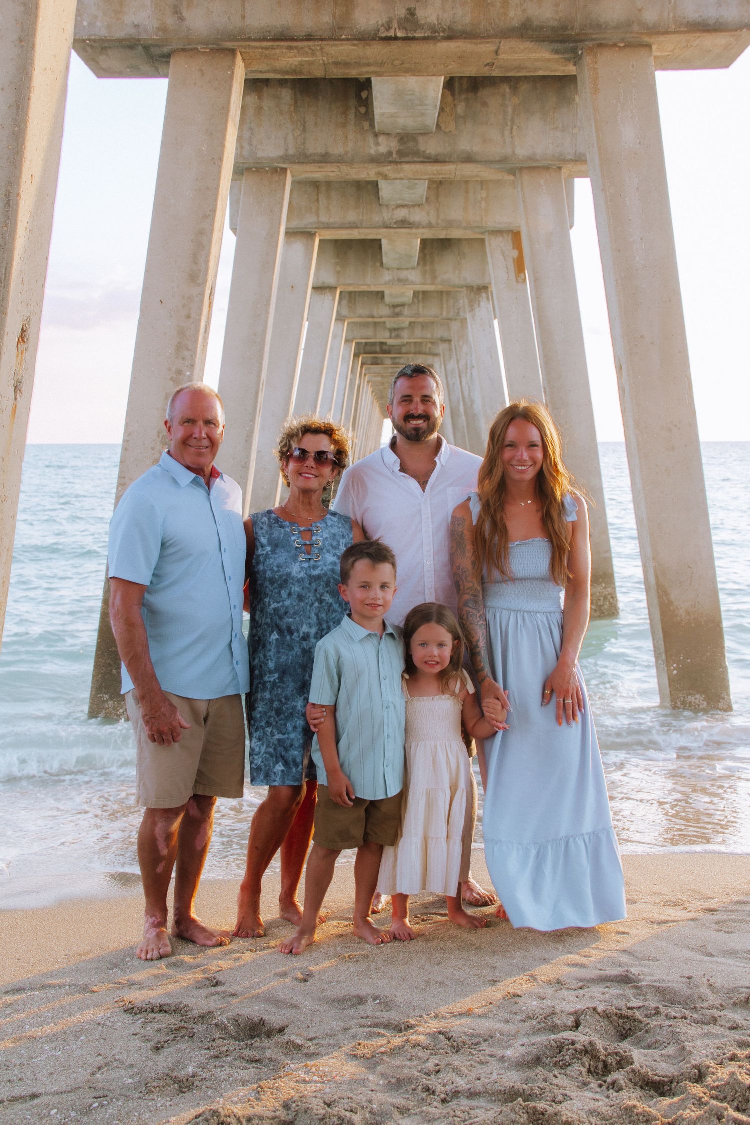 Multi-generational family of six poses together on sandy beach under a pier with ocean waves in the background.