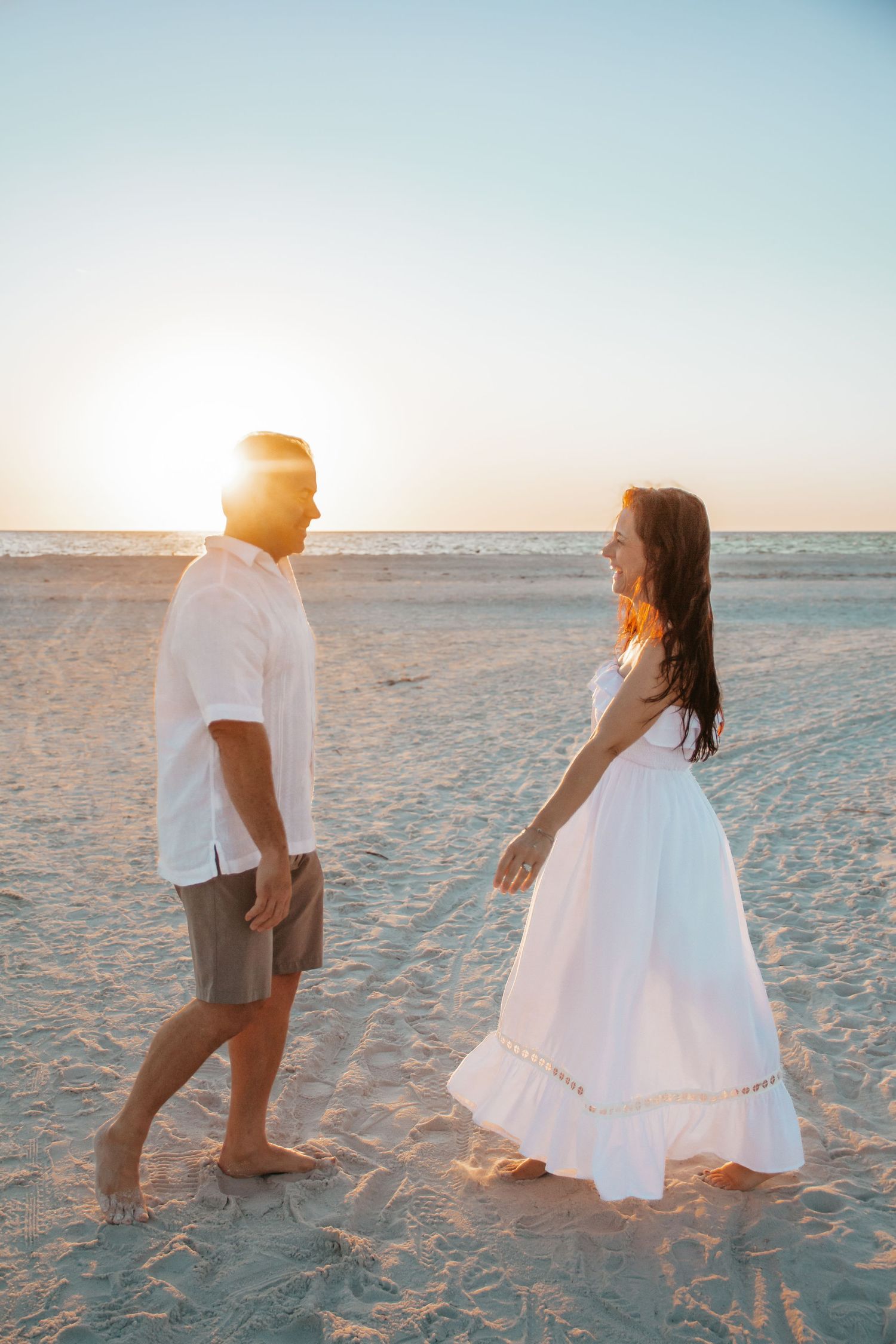 Couple in white outfits walking on a sandy beach at sunset, sharing a romantic moment by the ocean.