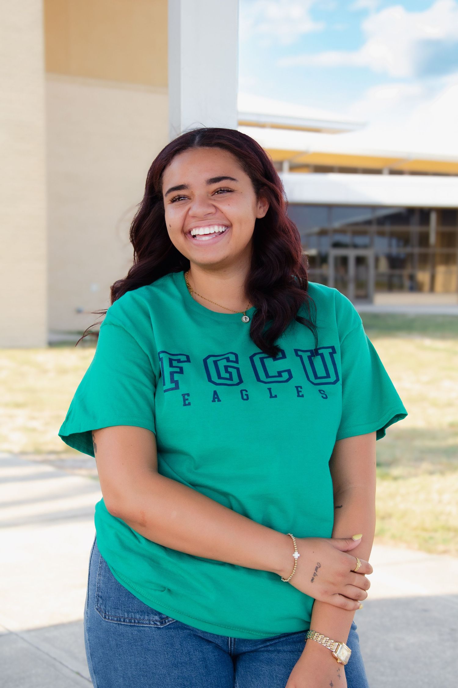 Smiling young woman wearing a green FGCU Eagles t-shirt and jeans, posing outdoors on a sunny day.