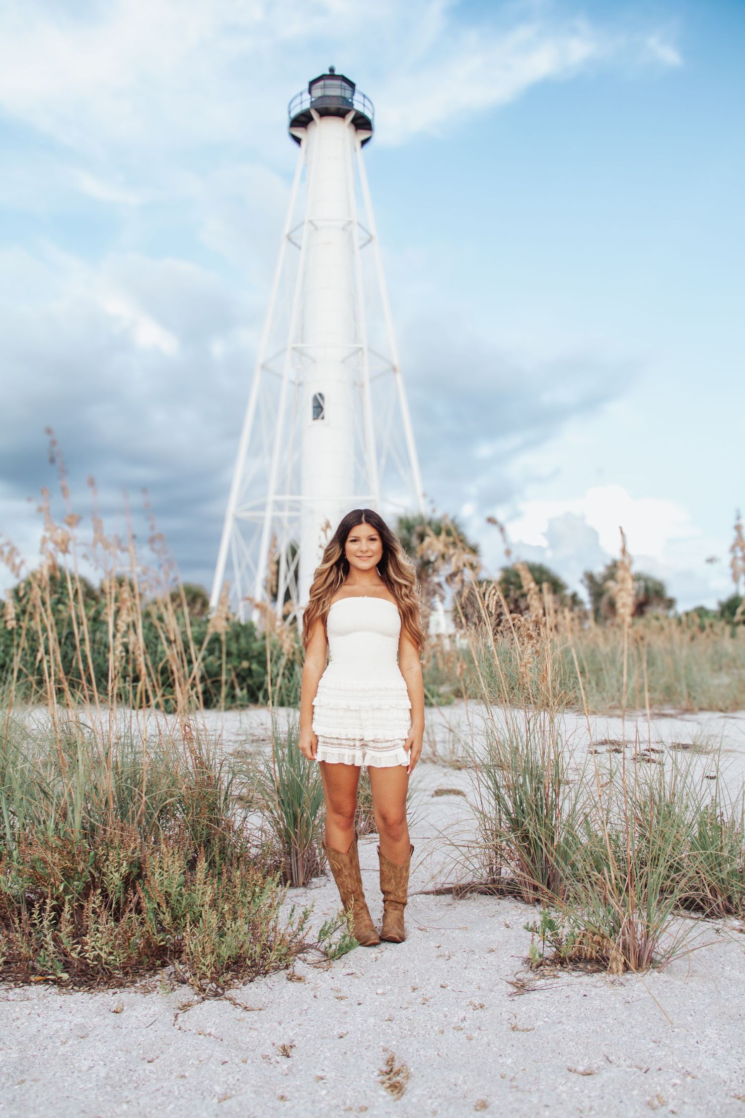 Woman in white dress and cowboy boots stands before a coastal lighthouse amid sea oats on sandy beach.