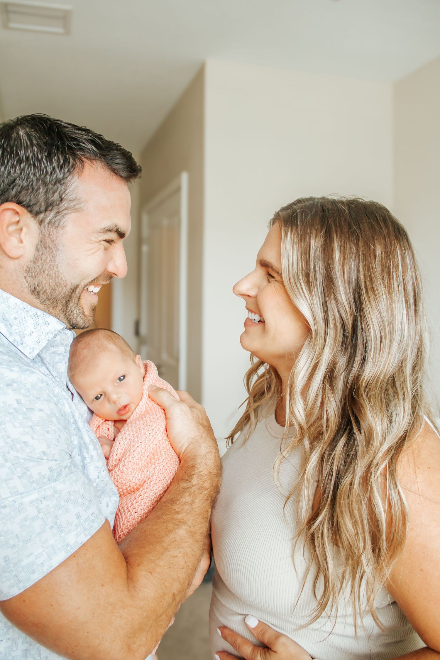 Happy couple smiling at each other while father holds newborn baby wrapped in pink swaddle blanket indoors.