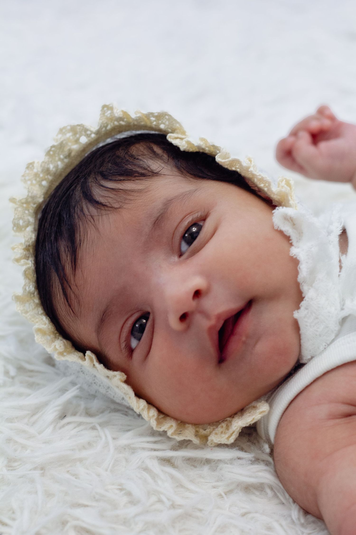Newborn baby wearing a delicate lace bonnet lying on a white fluffy blanket, looking up with bright eyes.