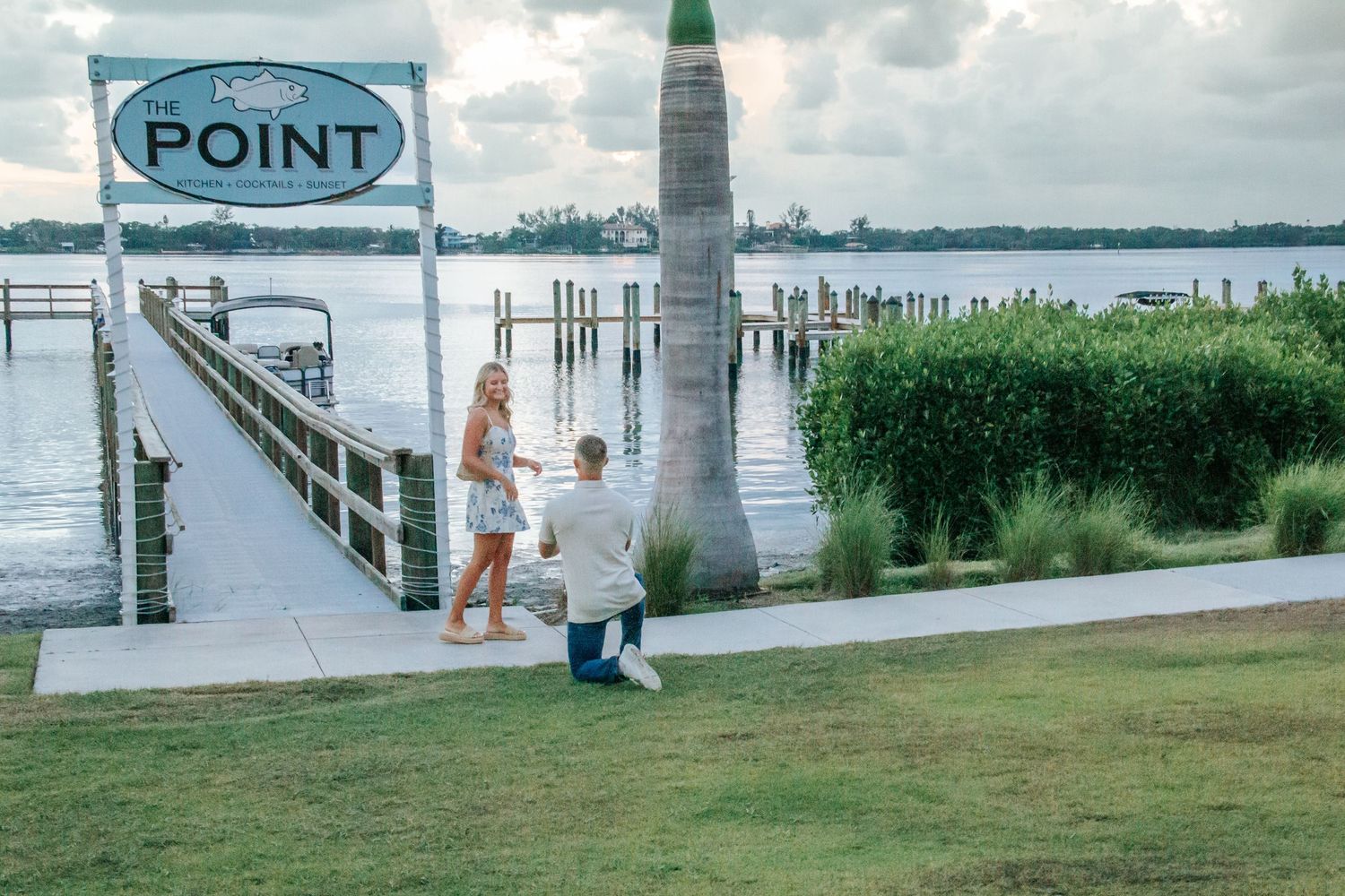 Man proposing to woman on one knee near The Point dock sign by a waterfront marina at sunset.