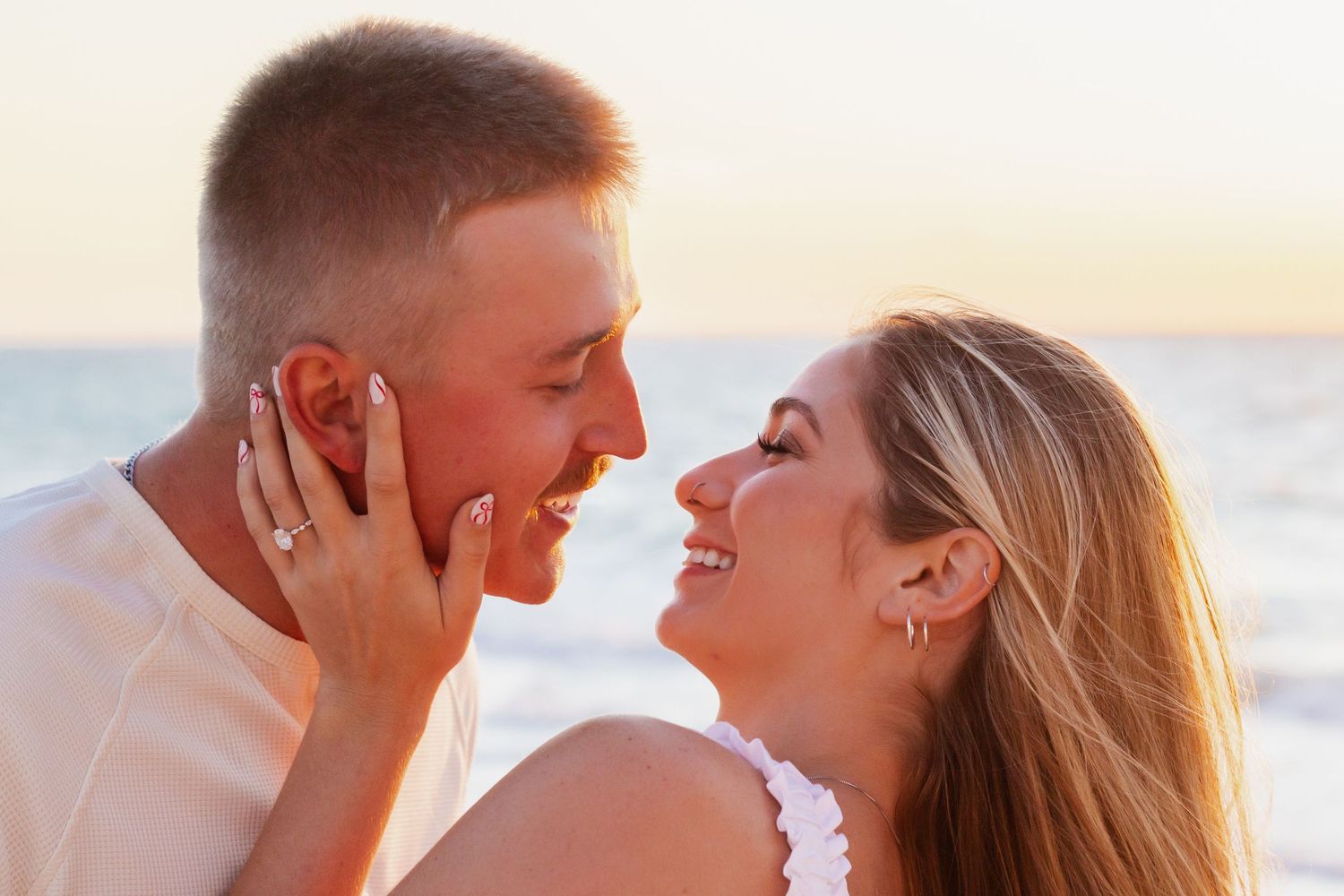 Happy couple sharing a romantic moment at sunset beach, woman wearing engagement ring, smiling face to face.