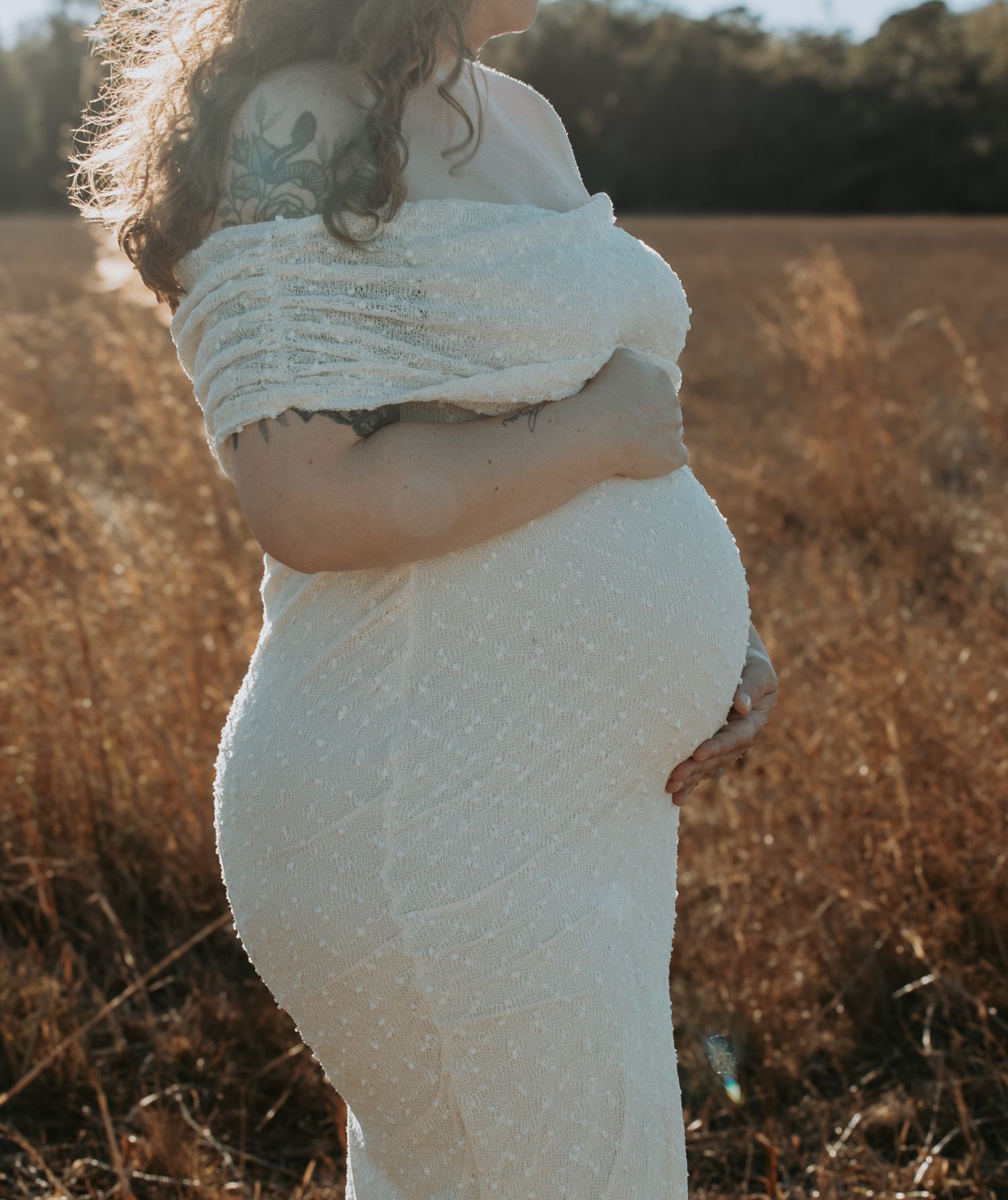 Pregnant woman in white lace off-shoulder gown standing in golden field, cradling baby bump during outdoor maternity photoshoot.