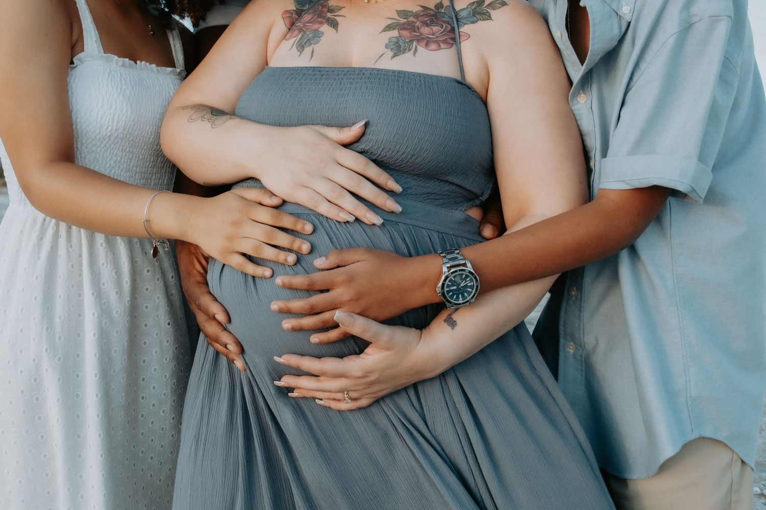 Pregnant woman in blue dress surrounded by loved ones gently placing hands on her baby bump.