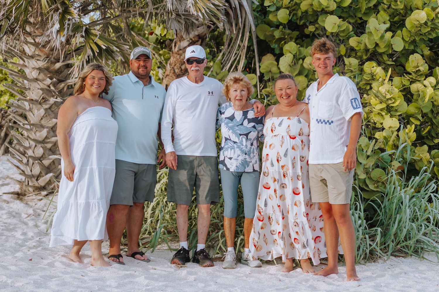 Family of six posing together on a sandy beach surrounded by tropical trees on a sunny day.