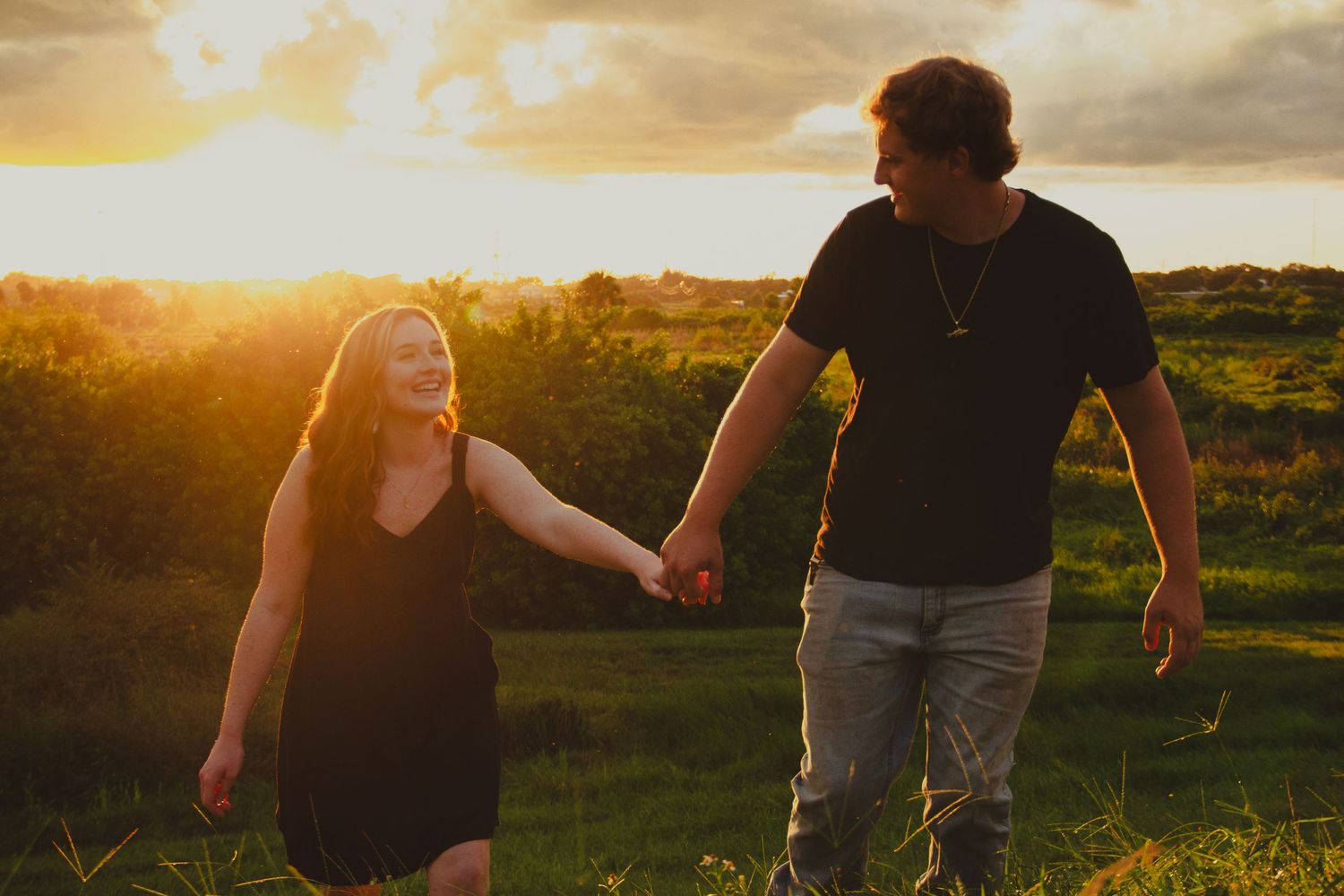 Couple holding hands walking through a green field during a golden sunset.