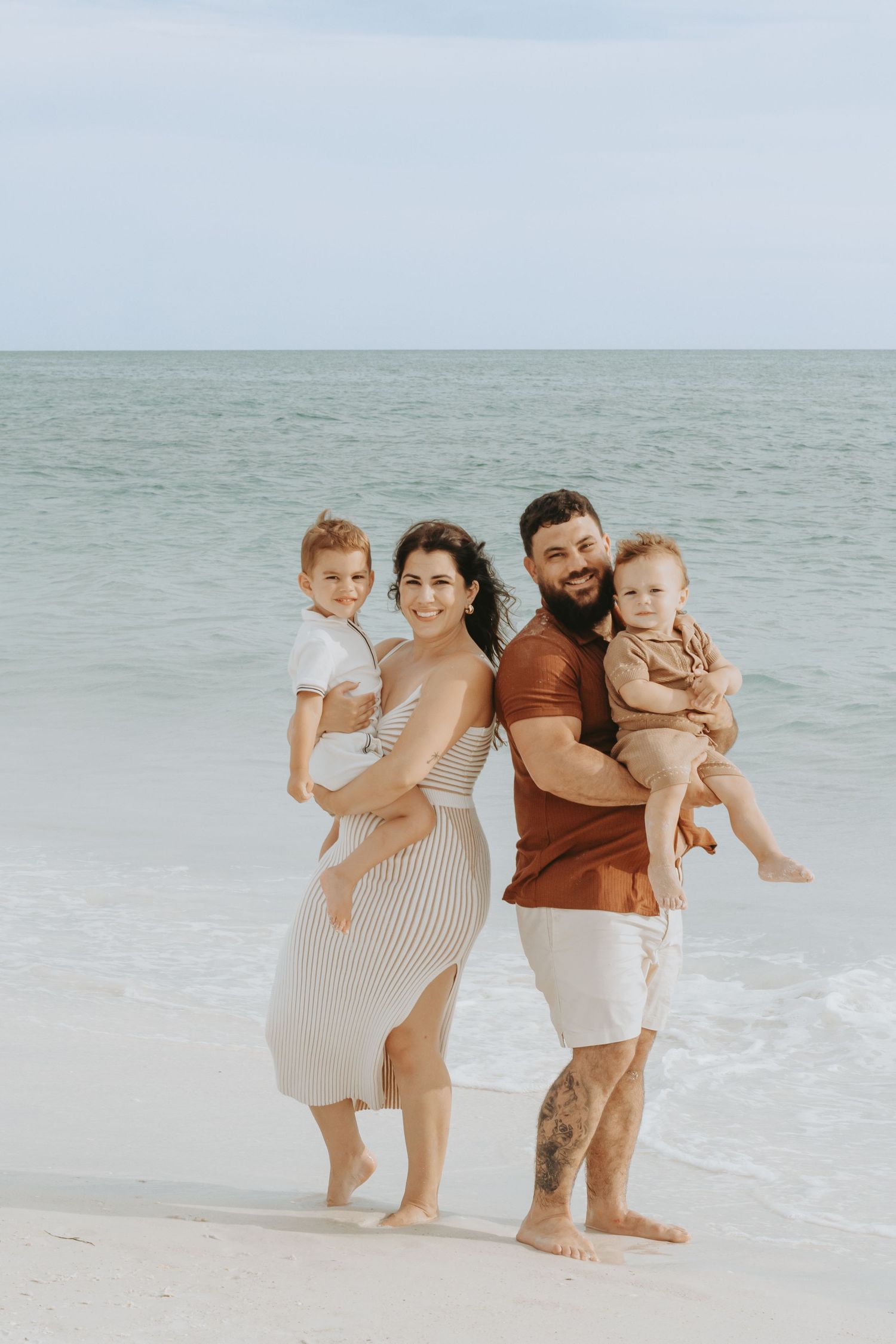 Happy family of four posing on a sunny beach, parents holding two young children with ocean waves in the background.