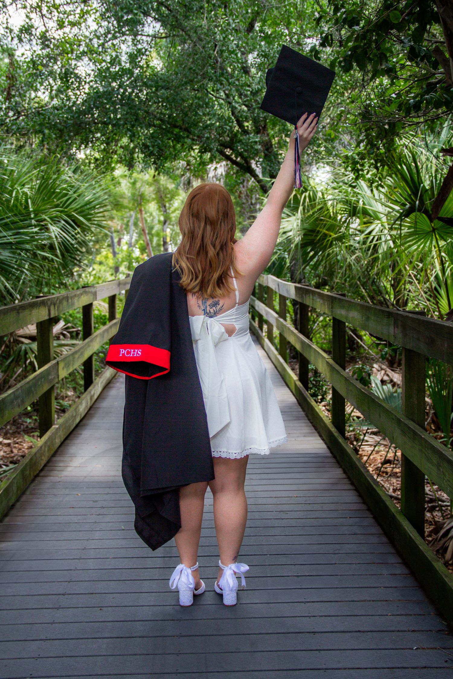 Graduate in white dress raises cap triumphantly on wooden bridge path surrounded by lush green tropical foliage.