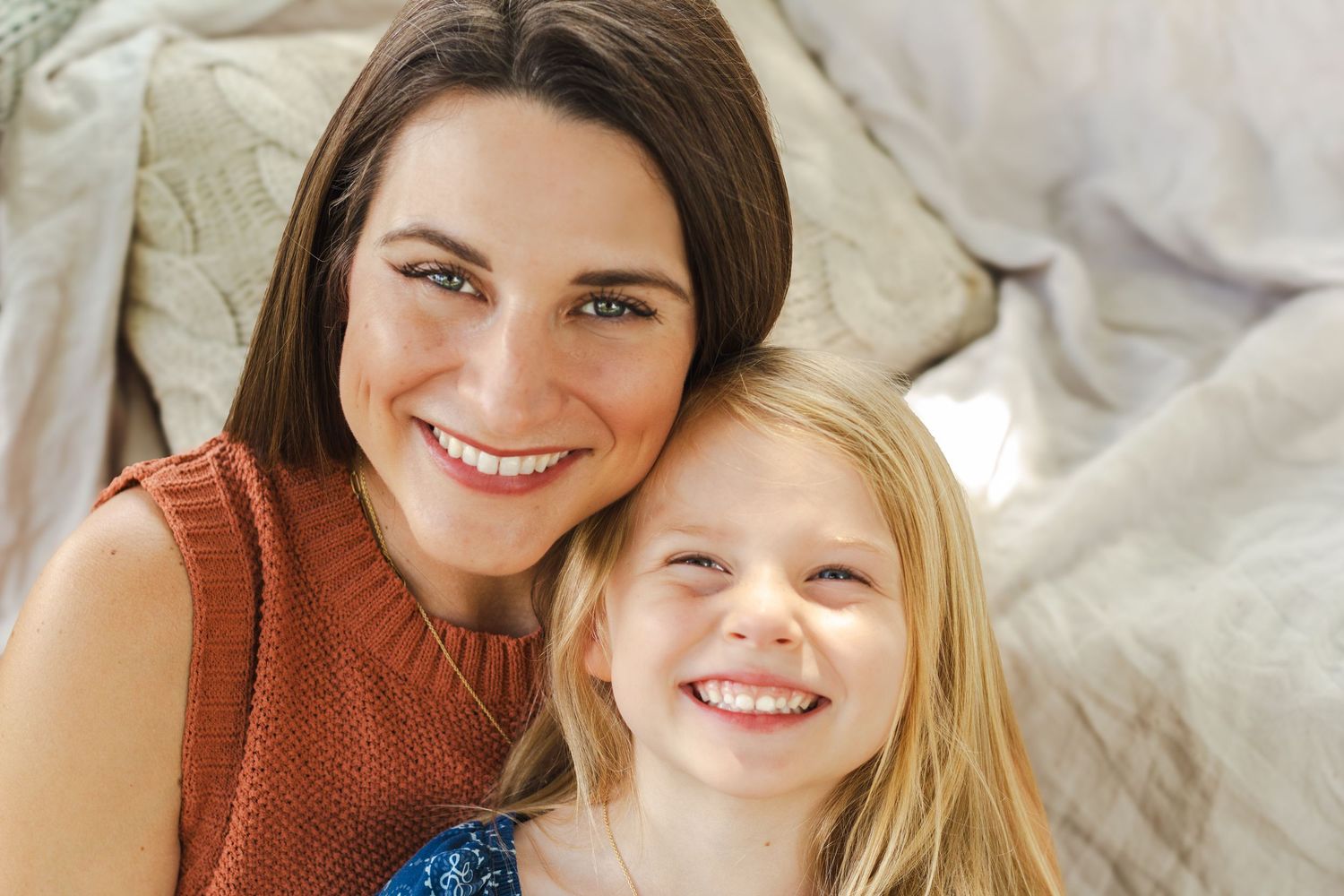 A smiling brunette woman and young blonde girl pose together cheek-to-cheek in a warm, cozy indoor setting.