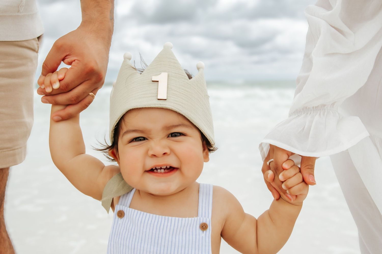 Toddler in a white beanie hat holding hands with adults while walking on a sunny day.