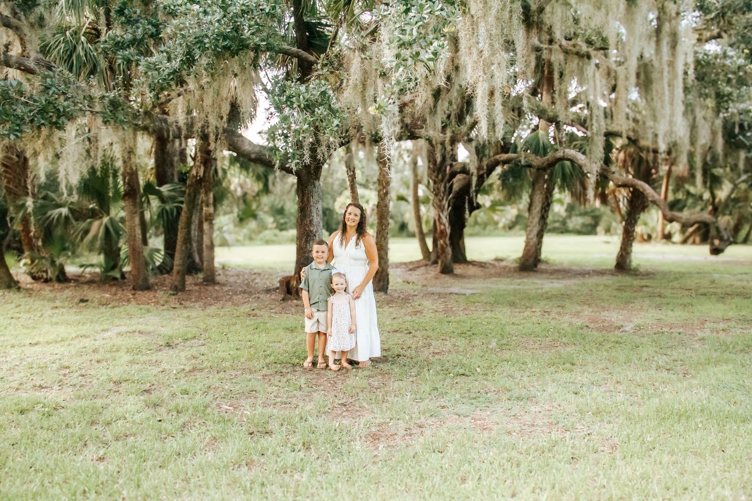 Three figures stand among moss-draped oak trees on a grassy lawn in a serene outdoor setting.