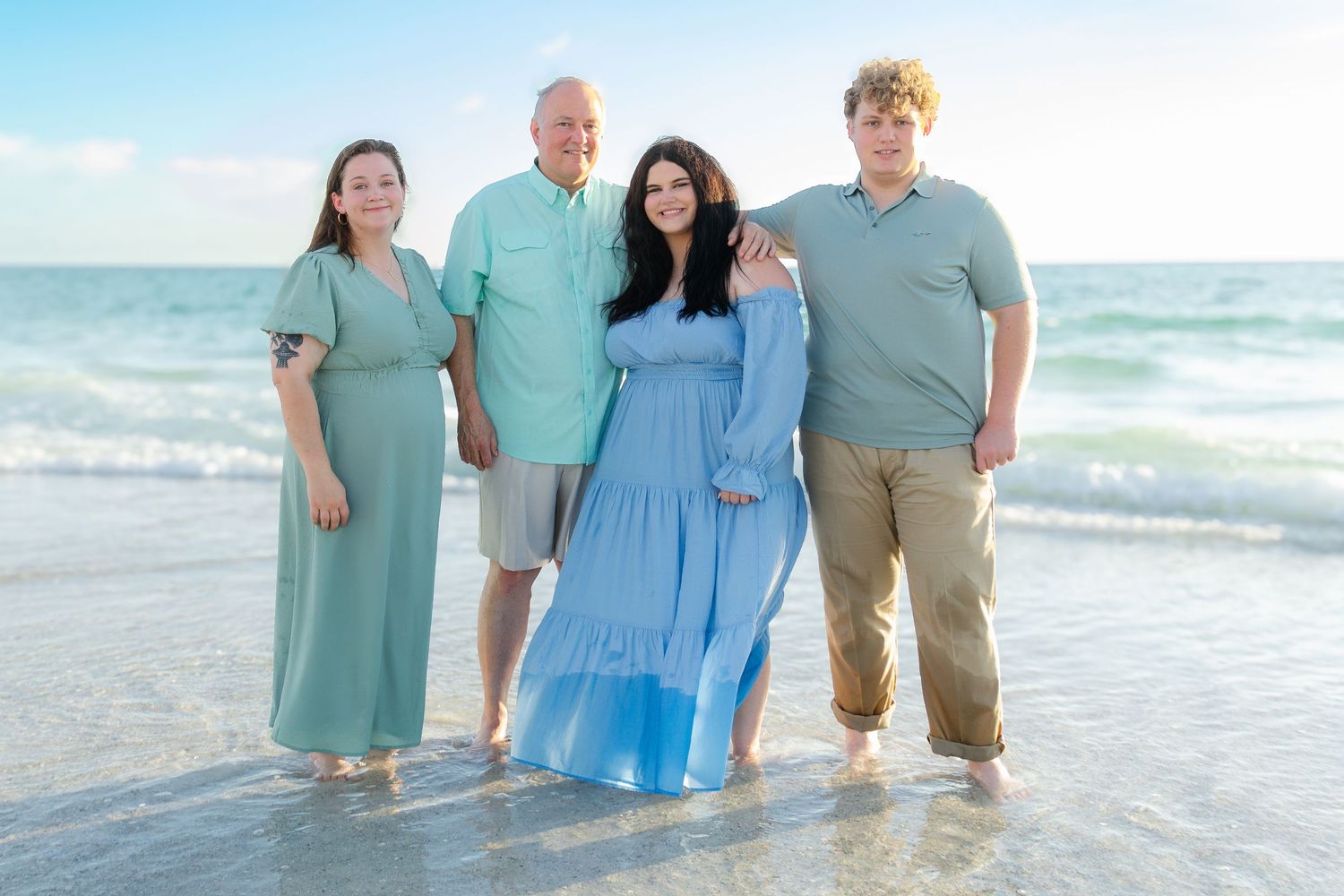 Family of four smiling together on a beach, wearing coordinating outfits in shades of blue and green, with ocean waves behind them.