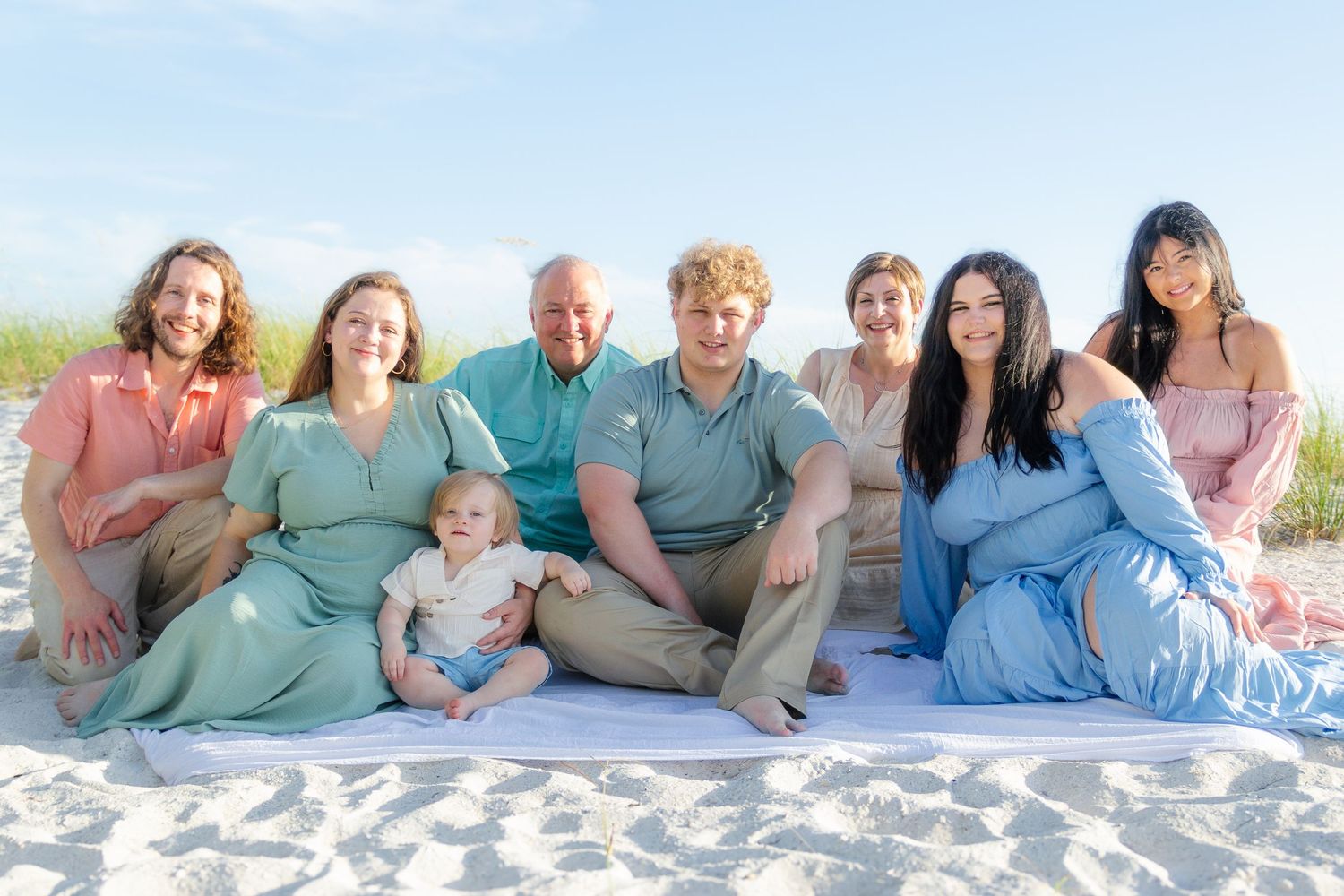Large family group photo on a sandy beach, smiling together in coordinated pastel outfits with a baby seated in front.