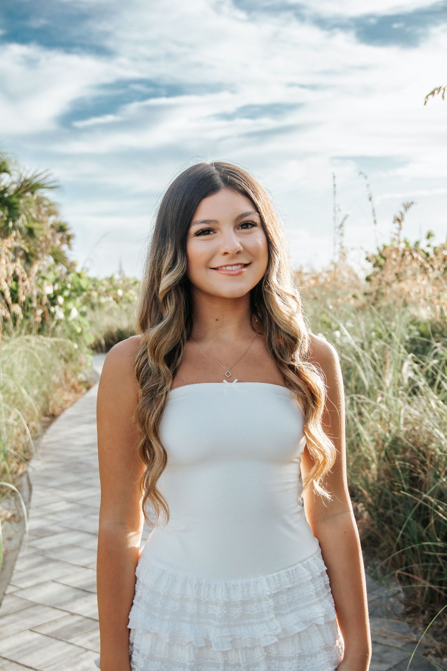 Young woman in white strapless ruffled dress smiling on a coastal boardwalk with sea grass and blue sky.