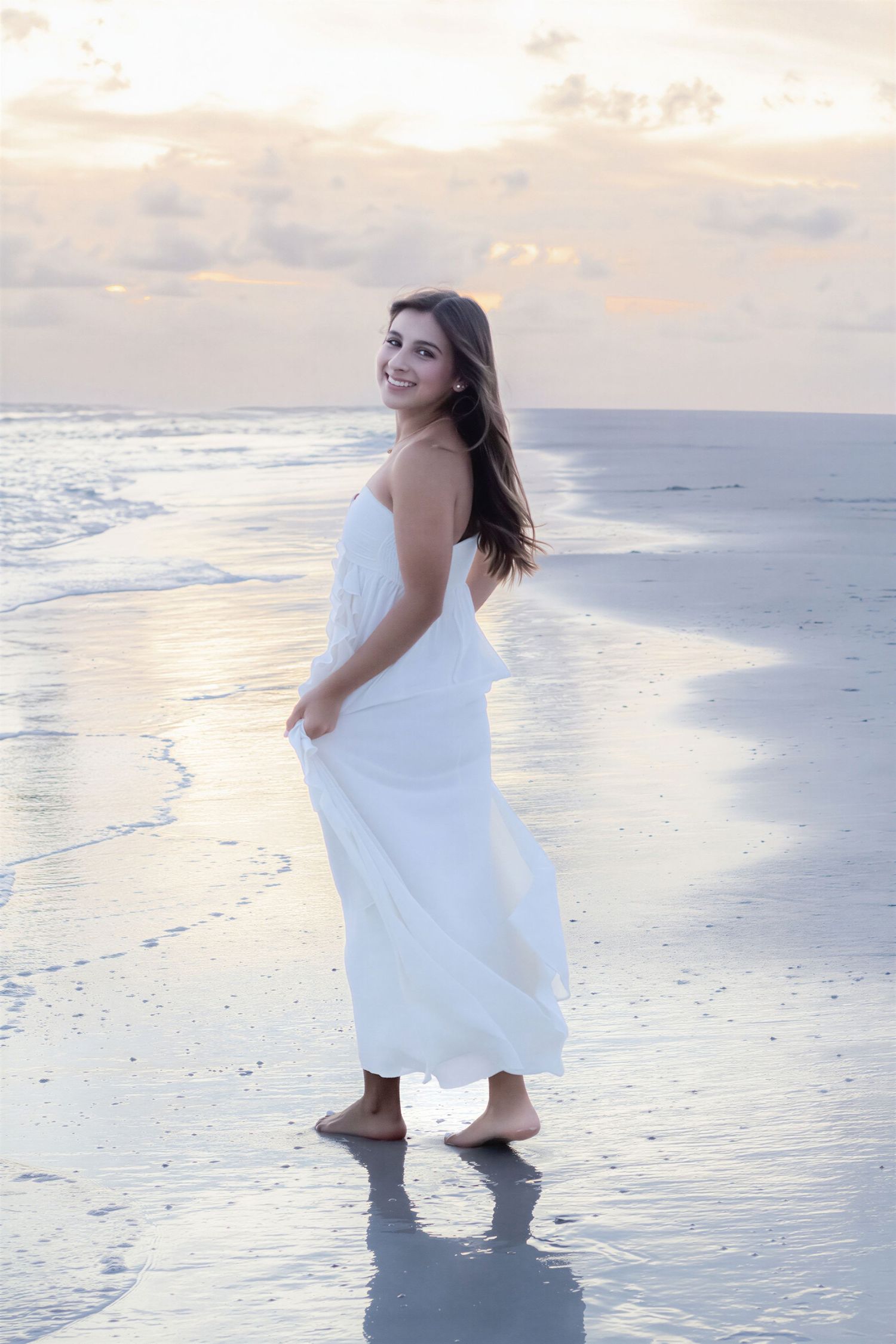 Young woman in white dress walking barefoot on beach at sunset, smiling over shoulder as waves wash ashore.