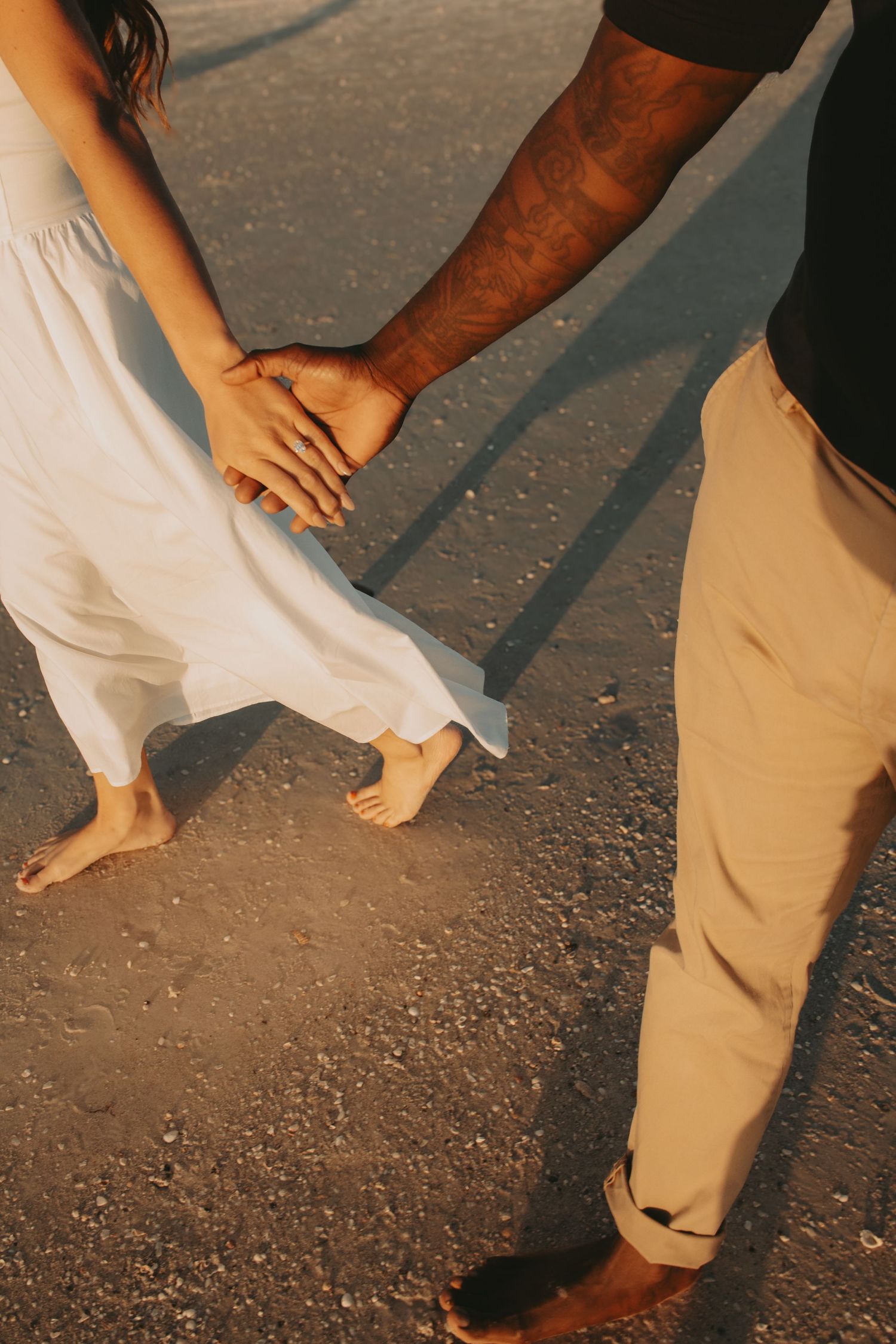 Couple holding hands barefoot on sandy beach at golden hour, woman in white dress, man with tattooed arm in beige pants.