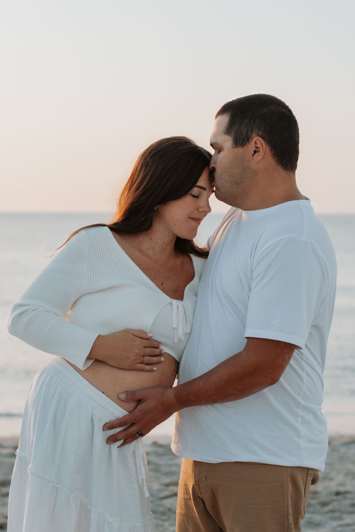 Couple embracing on beach at sunset, pregnant woman in white dress with partner in white shirt cradling her baby bump.
