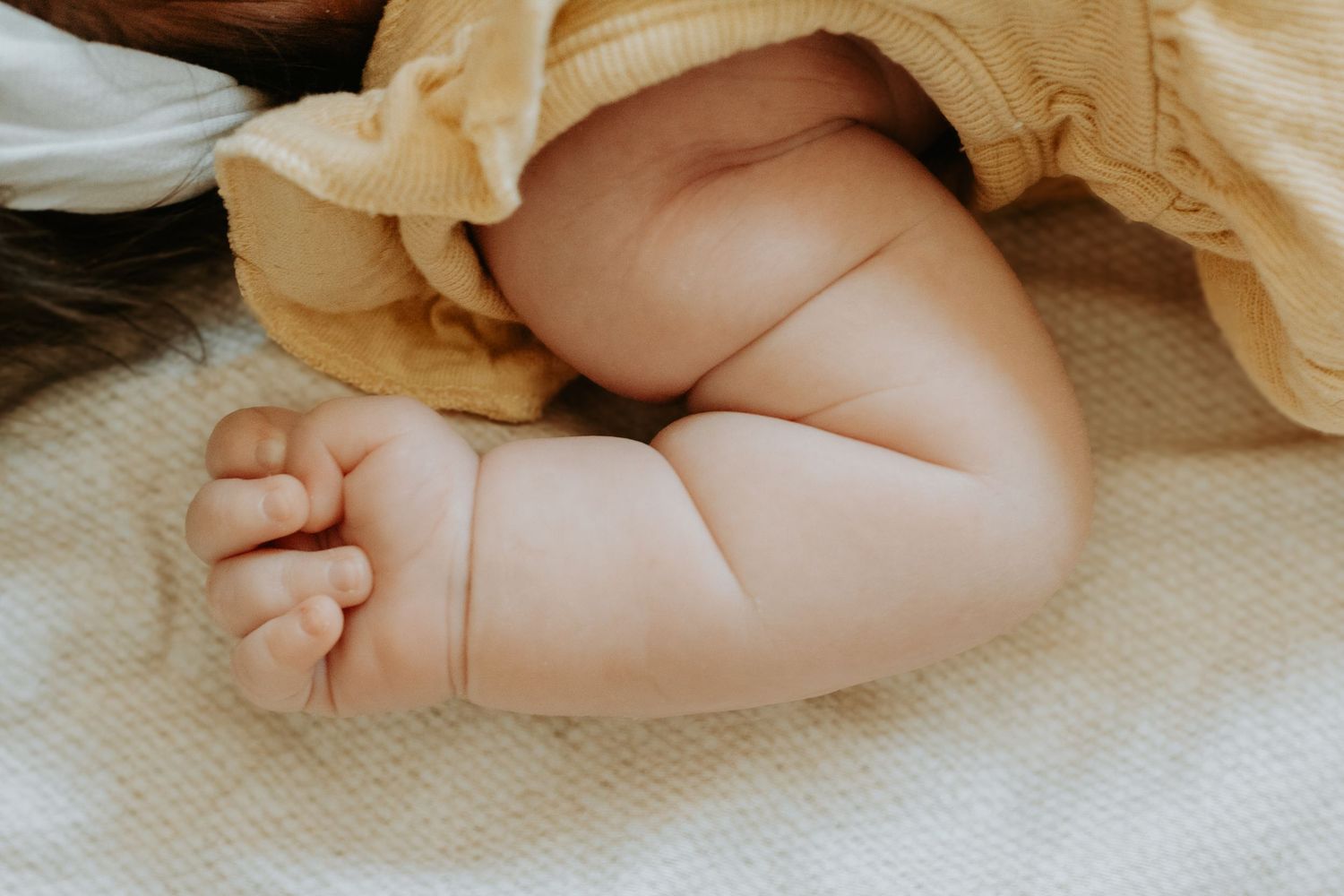 Close-up of a newborn baby's chubby arm and tiny hand resting on a white textured blanket in a yellow outfit.