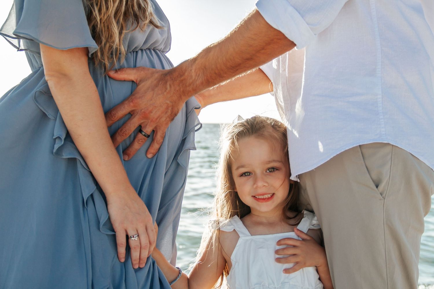 A young girl smiles between her parents near the ocean, with mom in a blue dress showing a baby bump.
