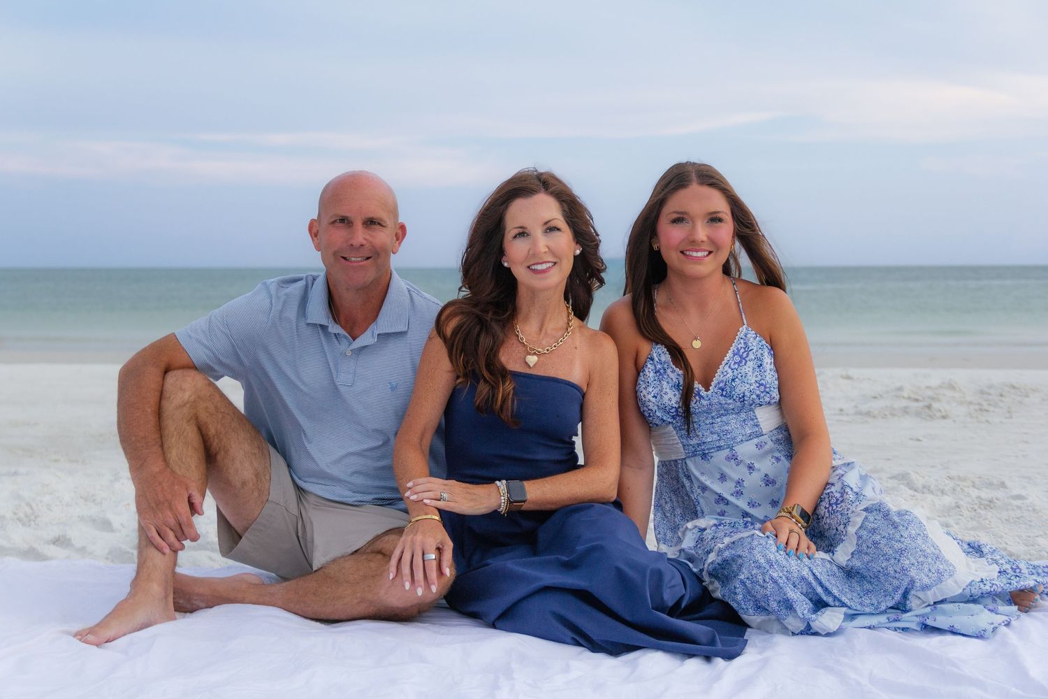 Family of three smiling together on a white sand beach with ocean waves in the background.