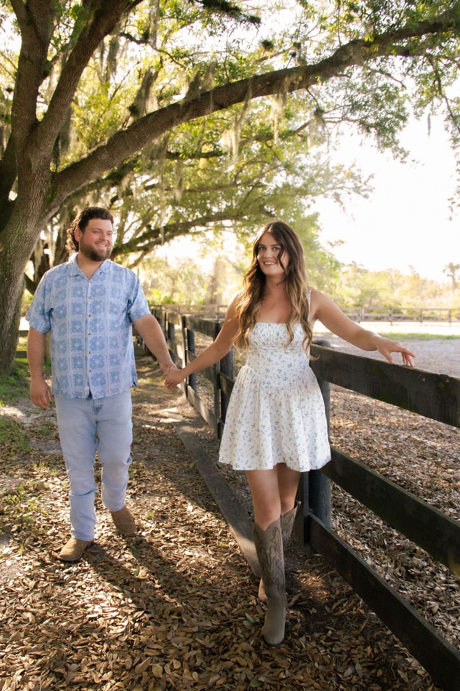 Couple holding hands walking along a wooden fence under oak trees, woman in white dress and cowboy boots.