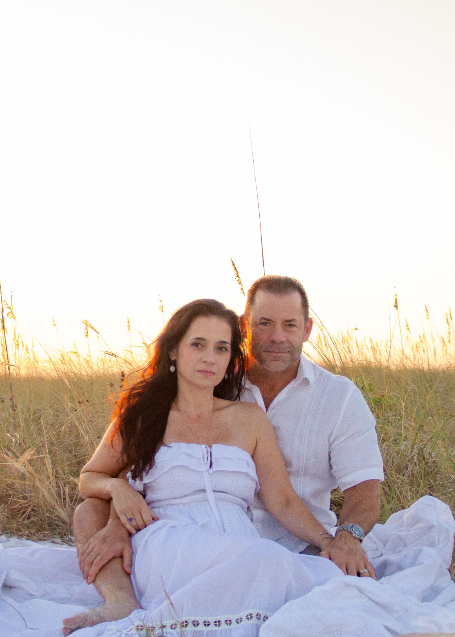 Couple in white outfits sitting together on a beach blanket among golden grass at sunset.