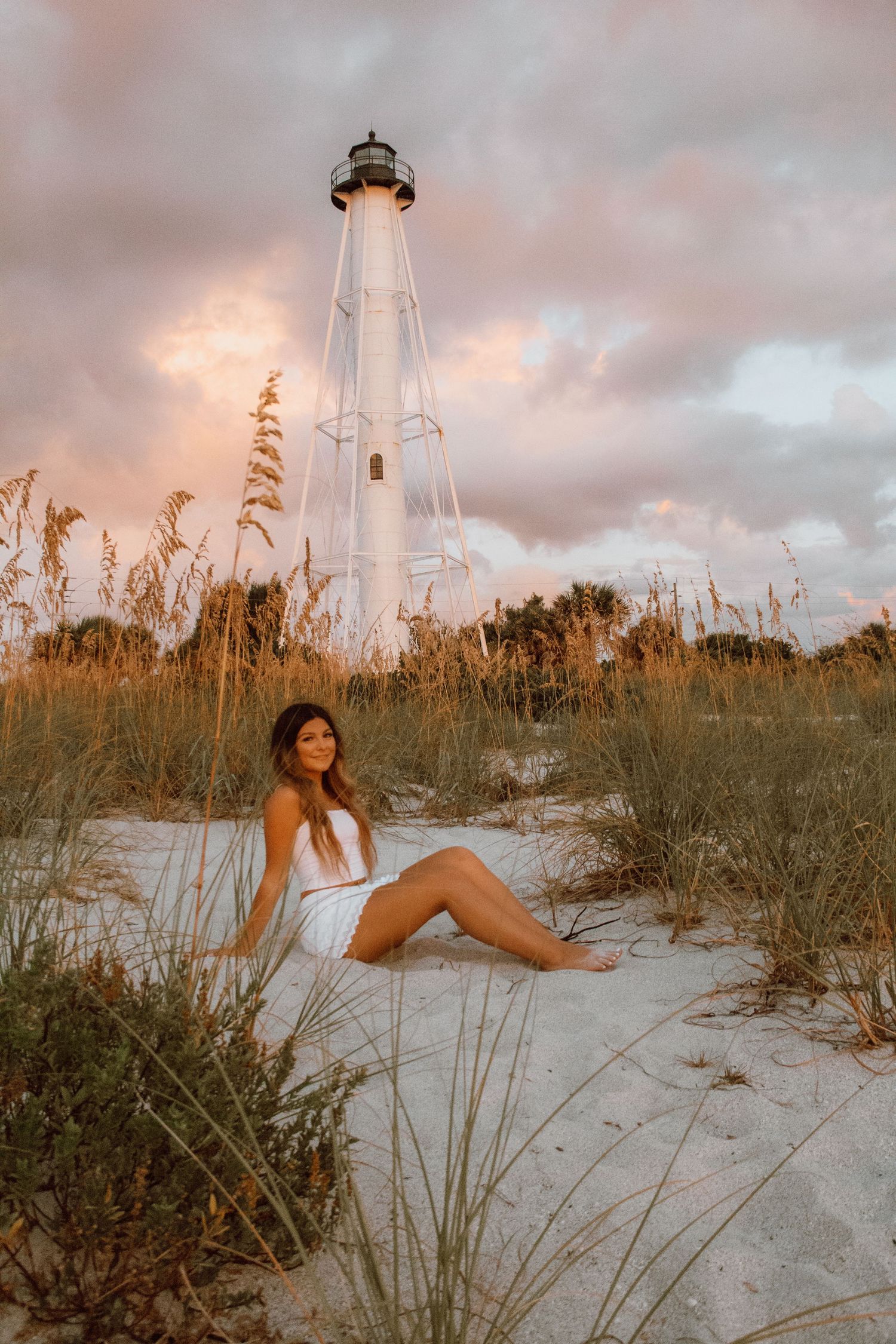 Woman in white dress sitting on sandy beach dunes near a tall white lighthouse at golden sunset hour.