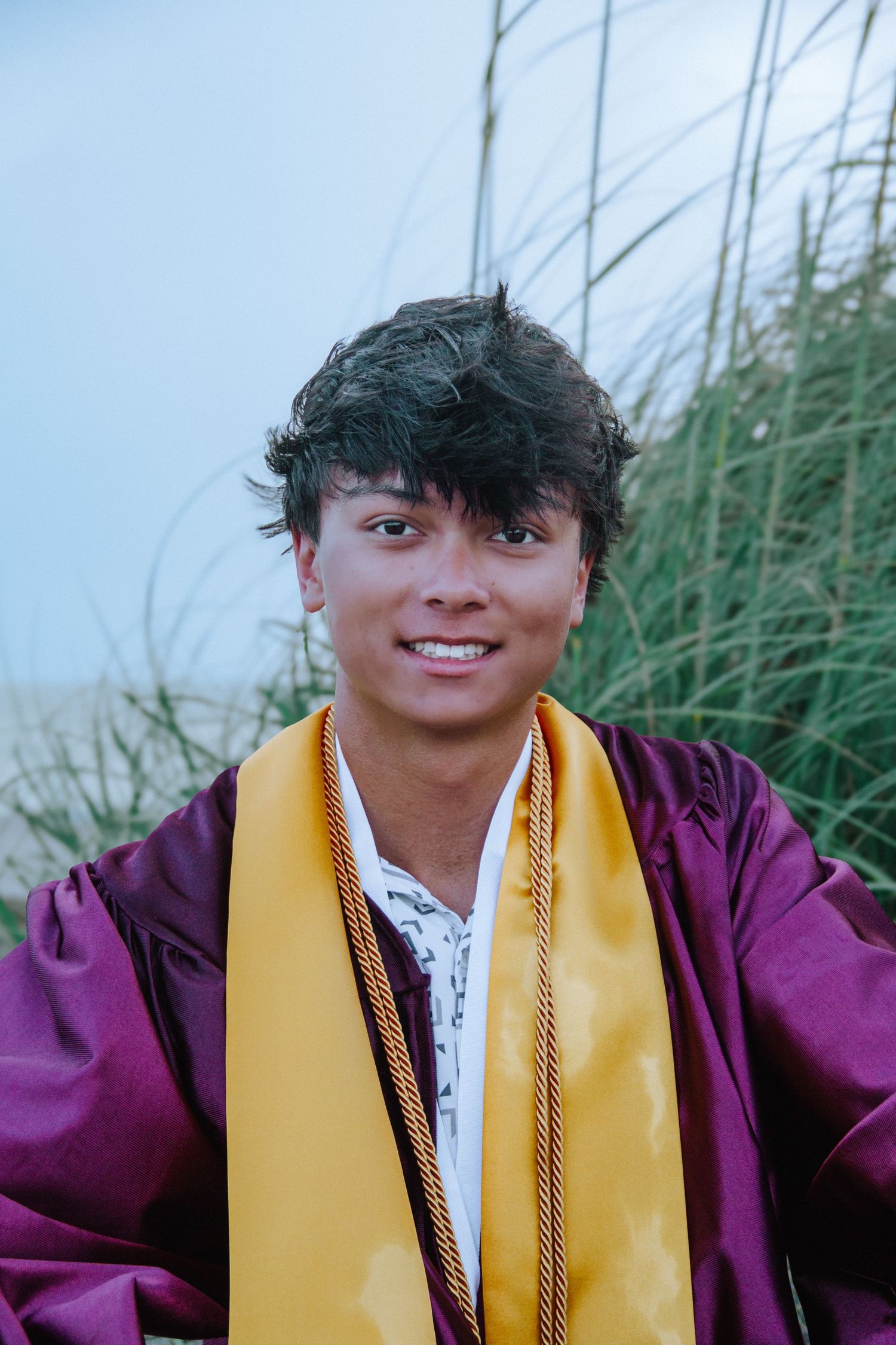 Smiling graduate in maroon cap and gown with gold stole poses outdoors near tall grass.